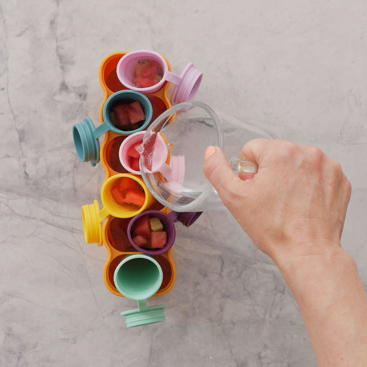 Hand holding a glass jug of water pouring it into silicone holders with fruit inside.