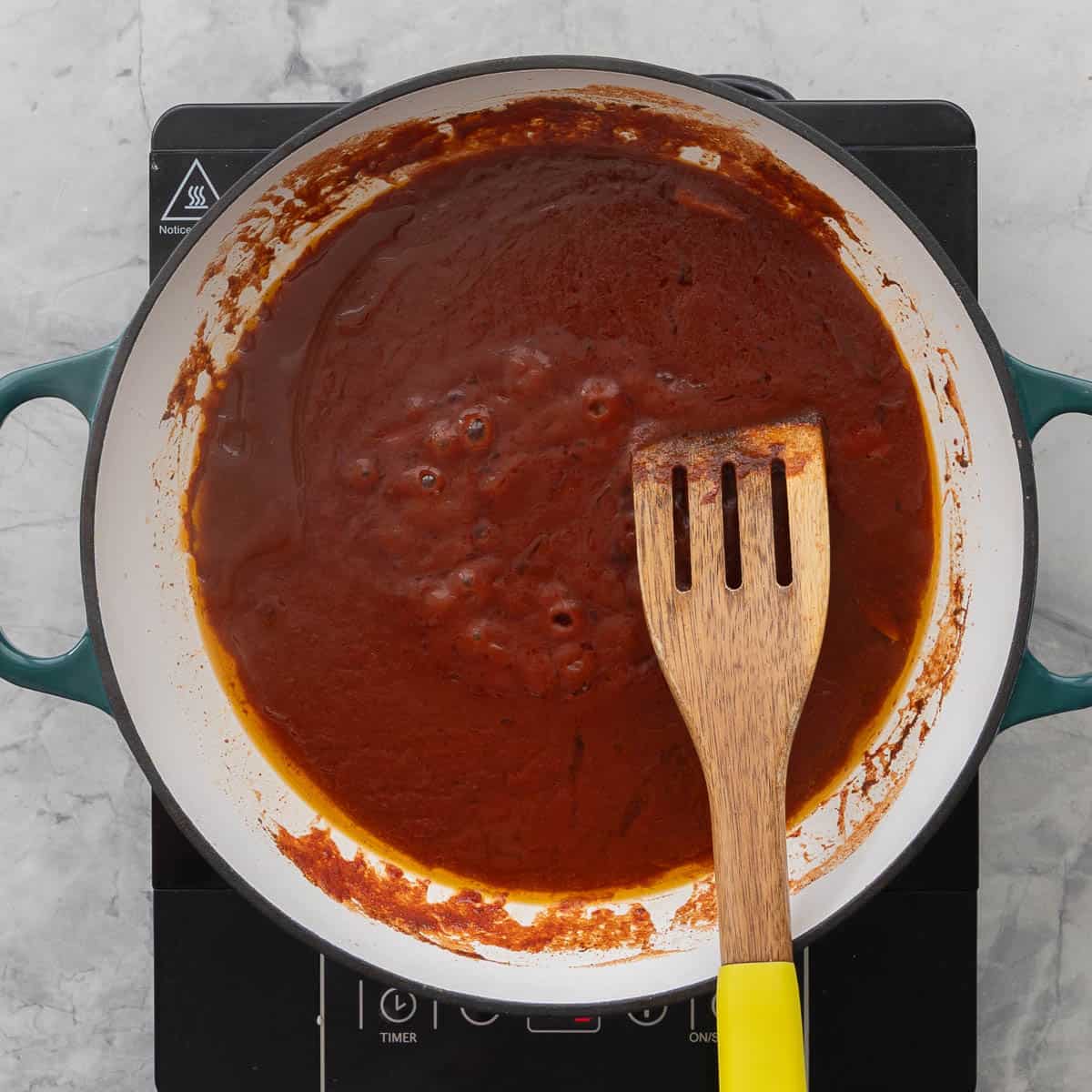 Wooden spatula stirring together bubbling tomato puree, cinnamon, maple syrup oil and garlic on stove top.