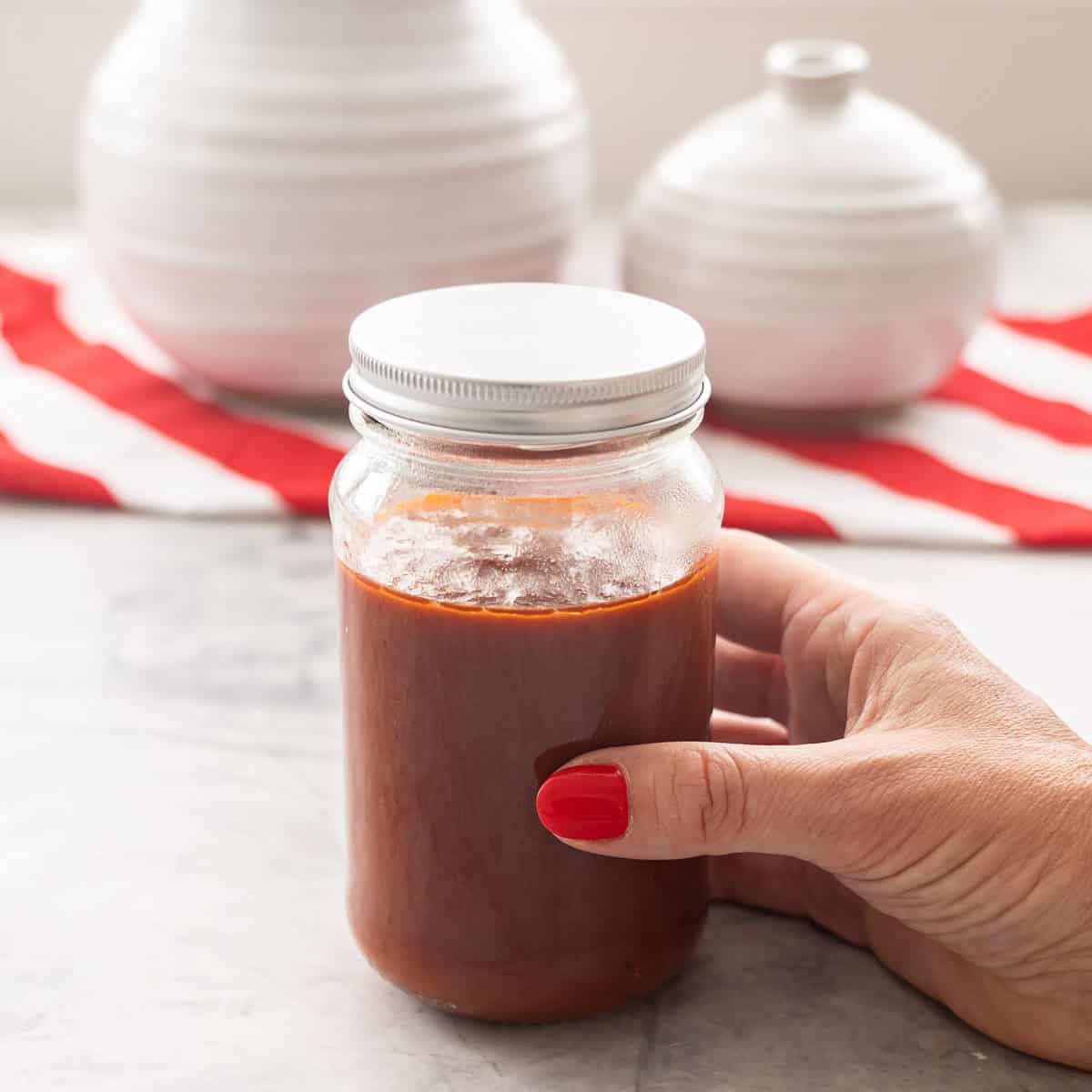 Hand holding a jar almost filled to the top with homemade ketchup inside. Red and white striped table runner on benchtop in background with white vases.