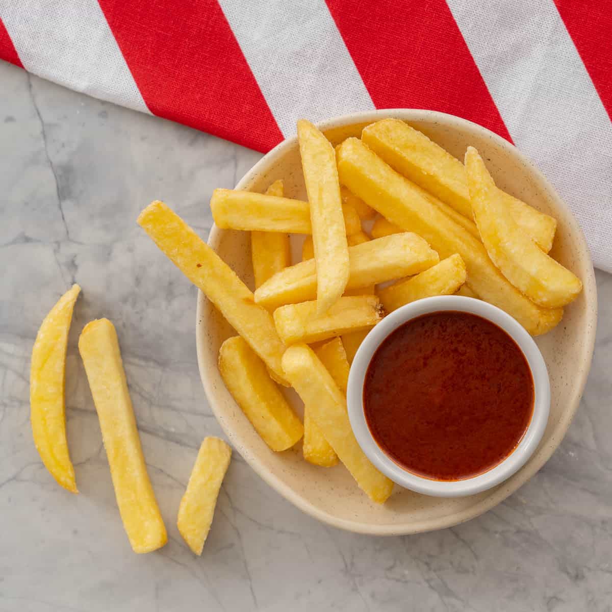 A bowl of fries with homemade ketchup in small ramekin inside bowl on benchtop with folded red and white stripped table cloth.