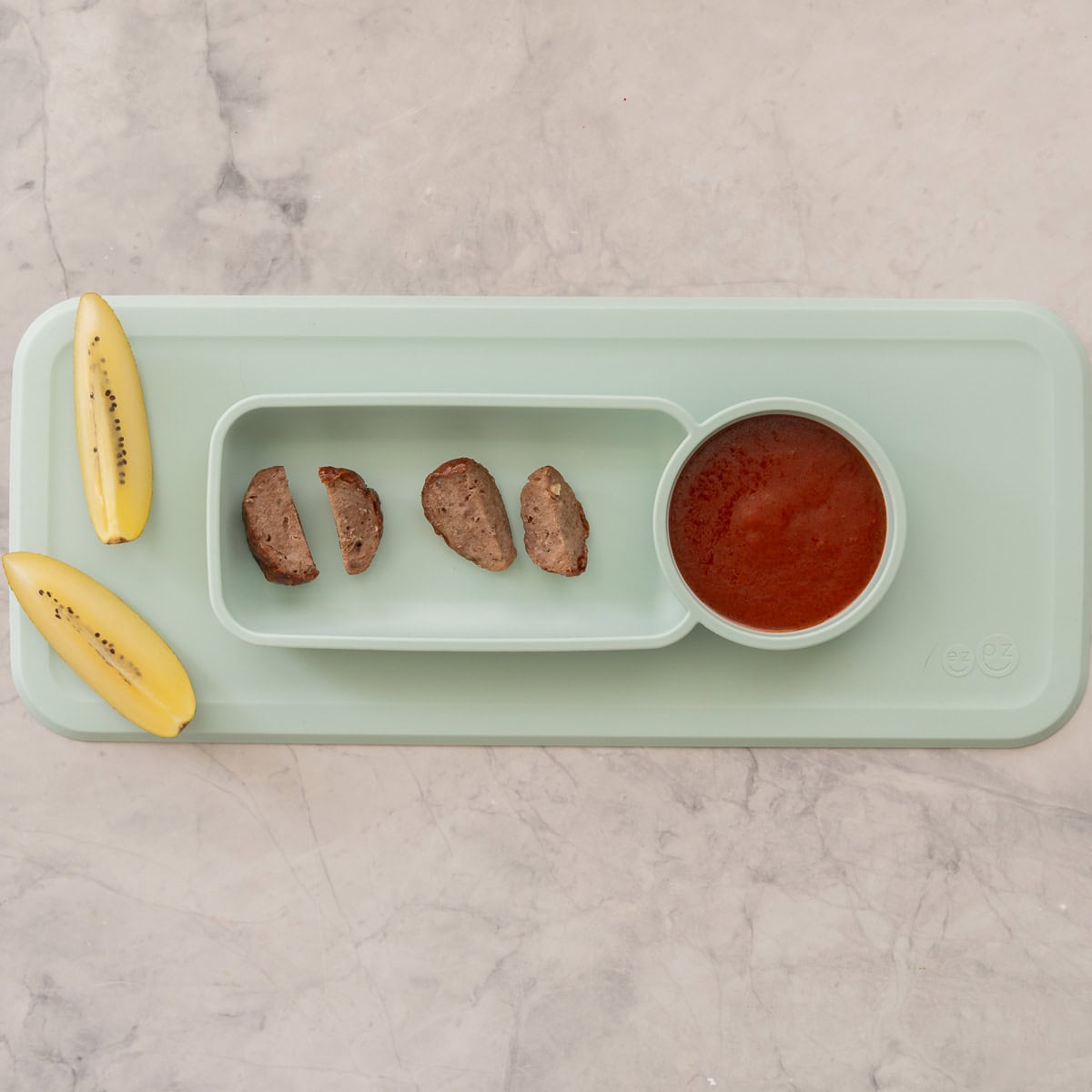 Silicone baby mat plate with four pieces of chopped up sausage, two slices of kiwifruit and homemade ketchup on the side.