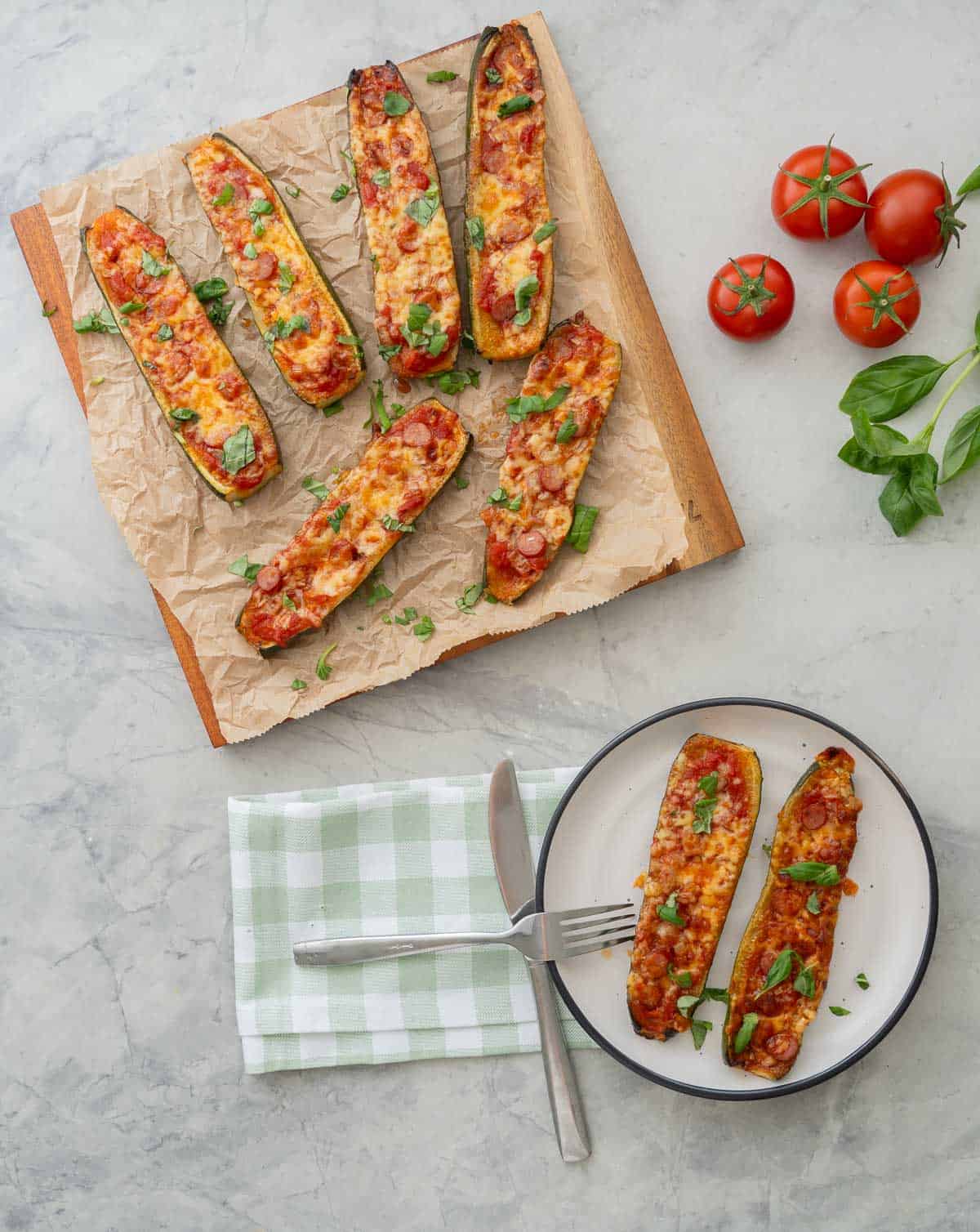 Cooked Zucchini Pizza boats laid on baking paper on wooden chopping board garnished with chopped basil, two zucchini boats on a dinner plate with knife and fork. Tomatoes, basil and a green checkered tea towel on benchtop.