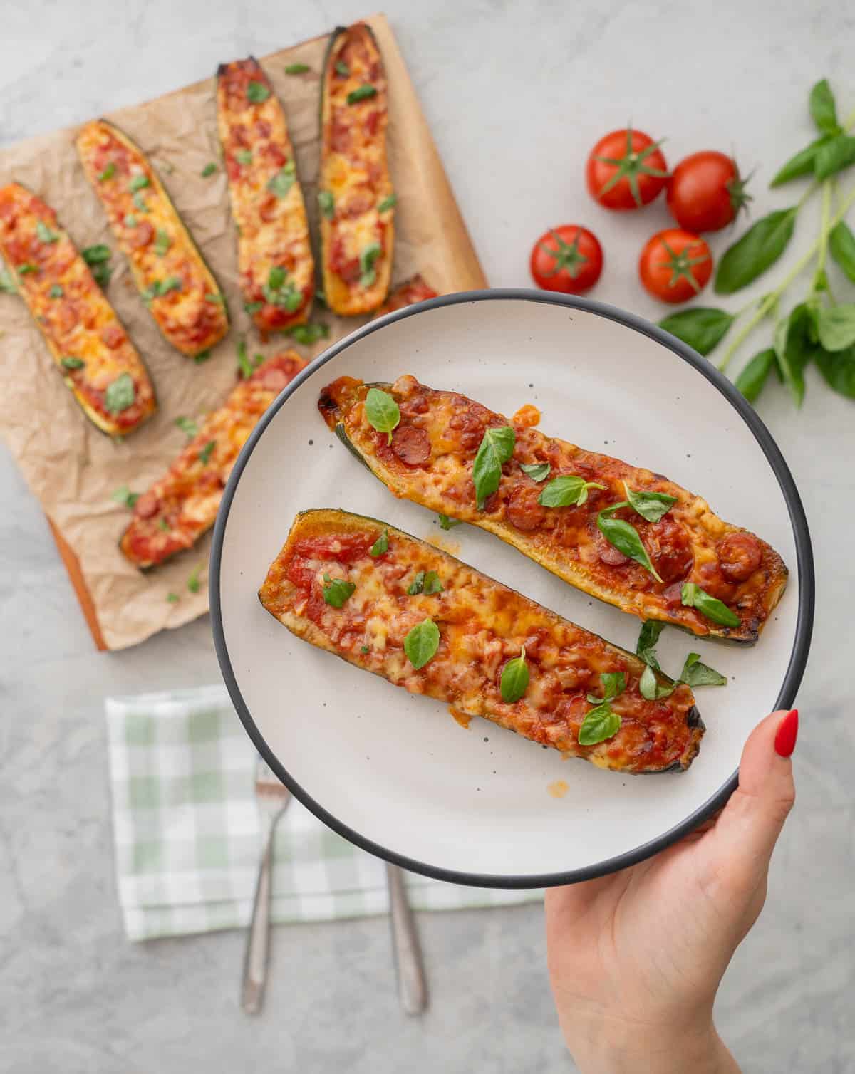 Hand holding a dinner plate with two zucchini boats. Cooked Zucchini Pizza boats laid on baking paper on wooden chopping board garnished with chopped basil, Tomatoes, basil and a green checkered tea towel on benchtop in background.