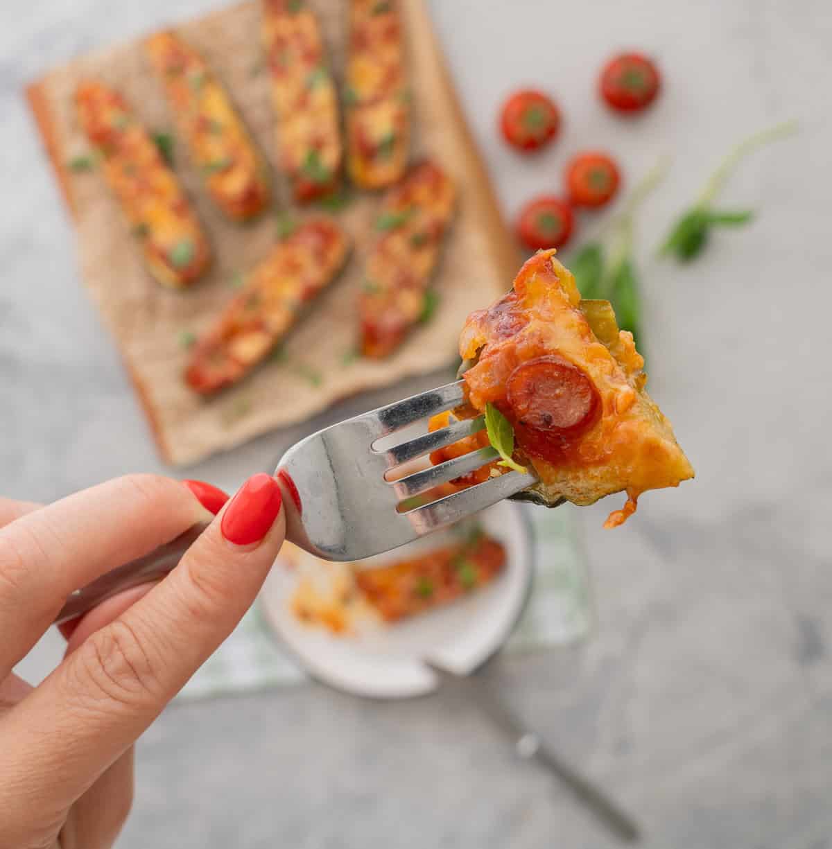 Hand holding a fork with a piece of zucchini pizza boat, Cooked Zucchini Pizza boats laid on baking paper on wooden chopping board garnished with chopped basil, Tomatoes, basil and a green checkered tea towel on benchtop in background.