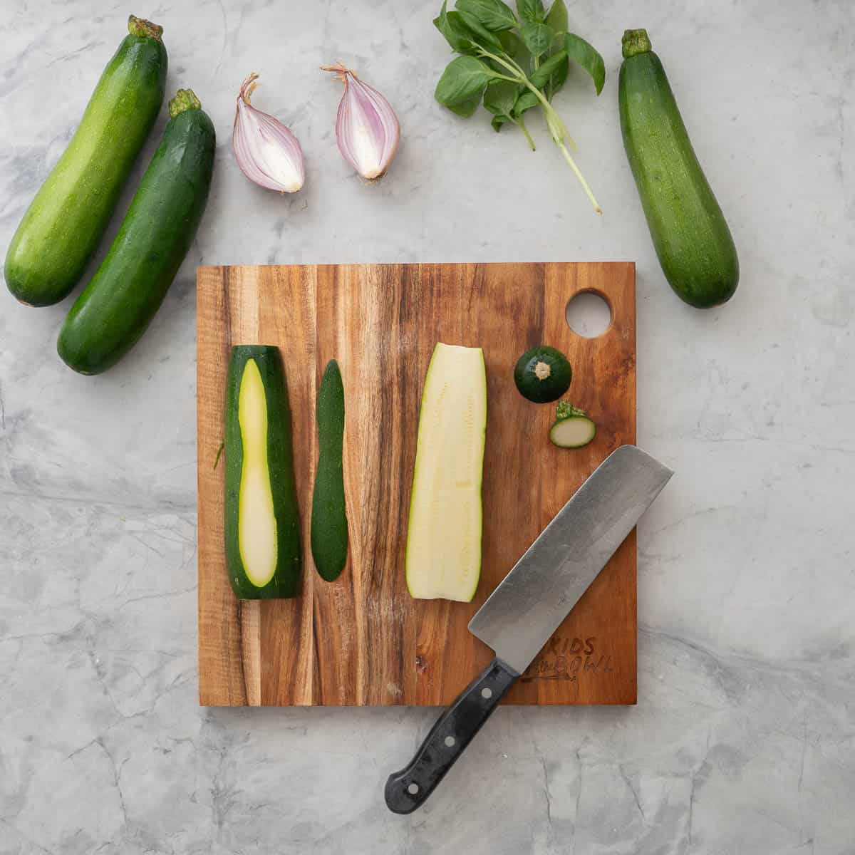 Zucchini cut in half lengthwise sitting on wooden chopping board, a small slice removed from the bottom of the half. Knife resting on chopping board, three zucchini, red onion and basil leaves on bench top.