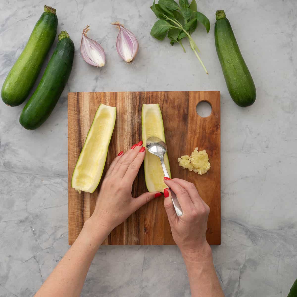 Zucchini cut in half lengthwise sitting on wooden chopping board, hand holding a spoon removing the flesh from the inside of zucchini. Three zucchini, red onion and basil leaves on bench top.