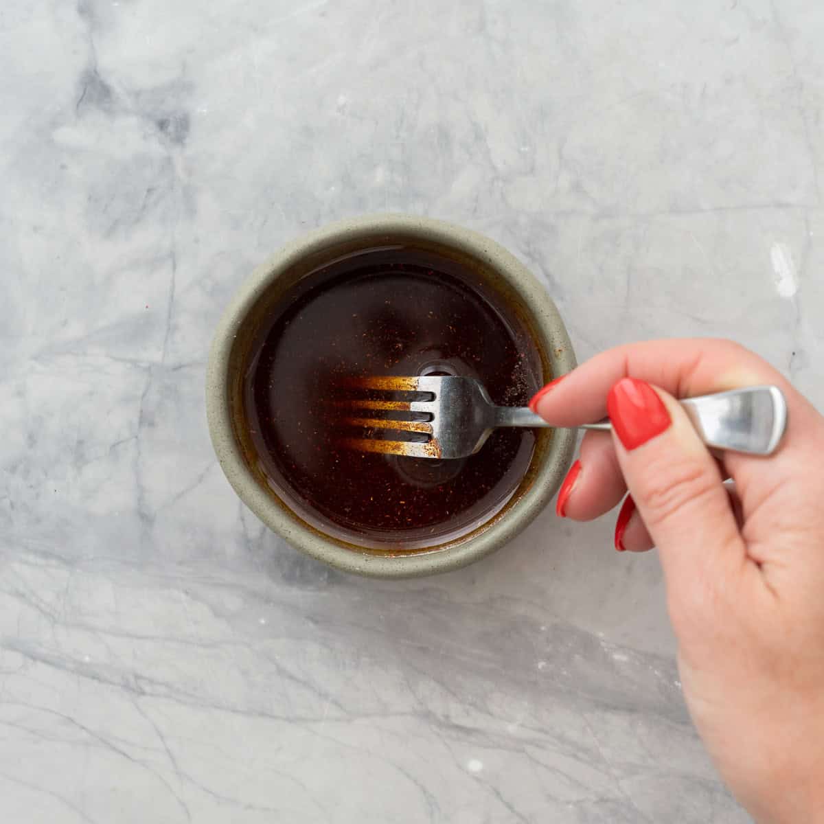 Hand holding a fork stirring together olive oil, paprika and garlic powder in a small ramekin on benchtop.