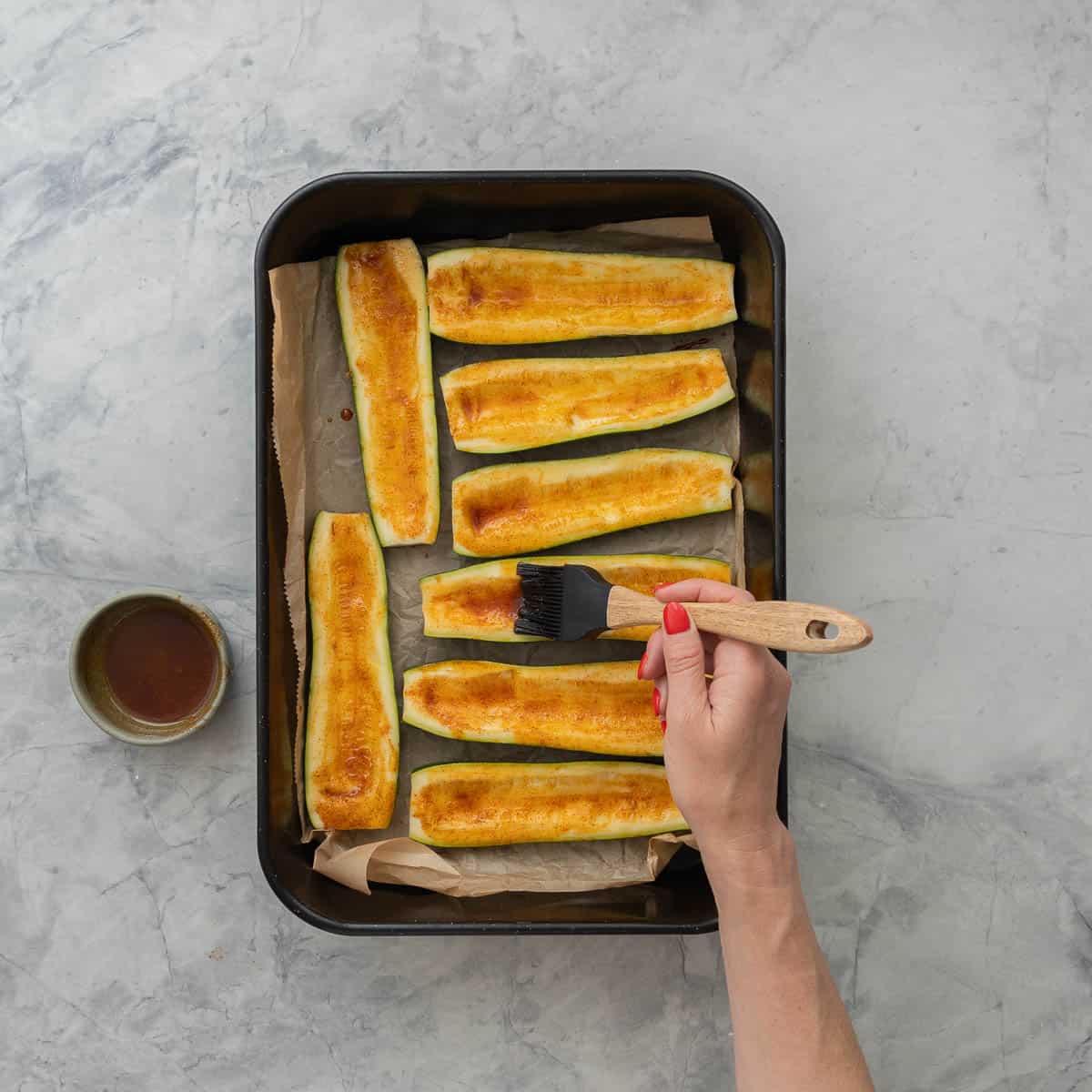 Eight hallowed out zucchini inside of a lined baking tray, hand brushing on Olive oil, paprika and garlic powder mixture.