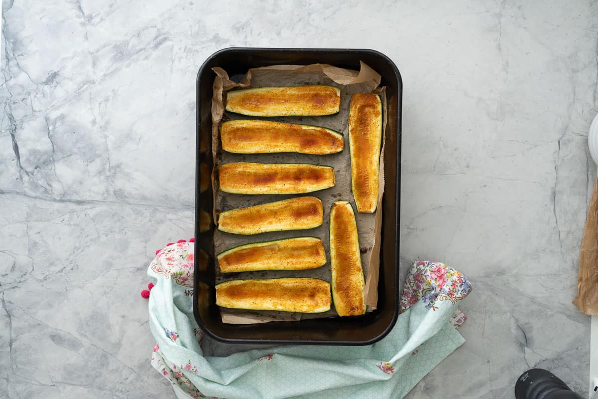 Cooked zucchini pizza boats inside a baking tray on bench top.