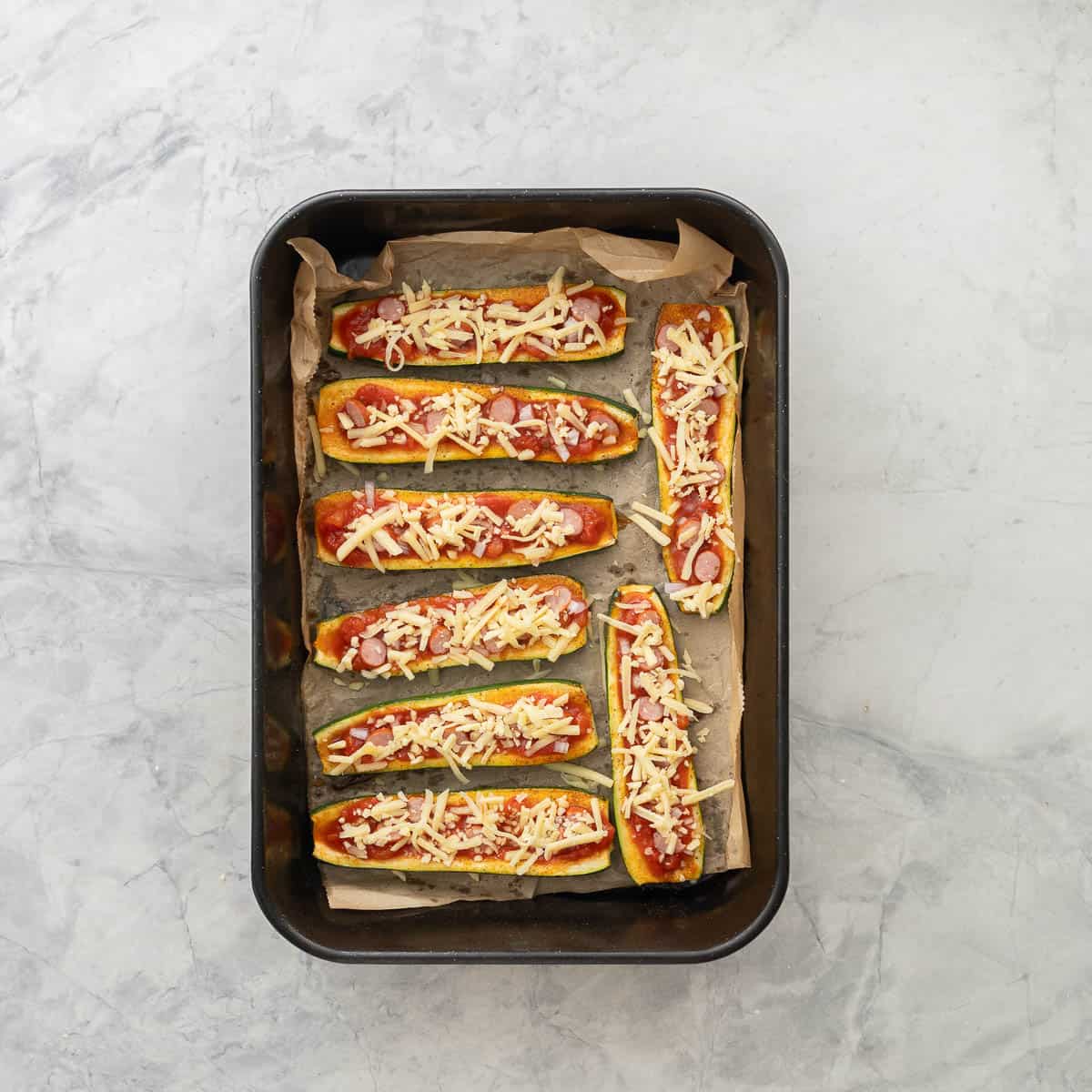 Cooked zucchini pizza boats in baking tray topped with Pizza Sauce, red onion, salami and mozzarella.