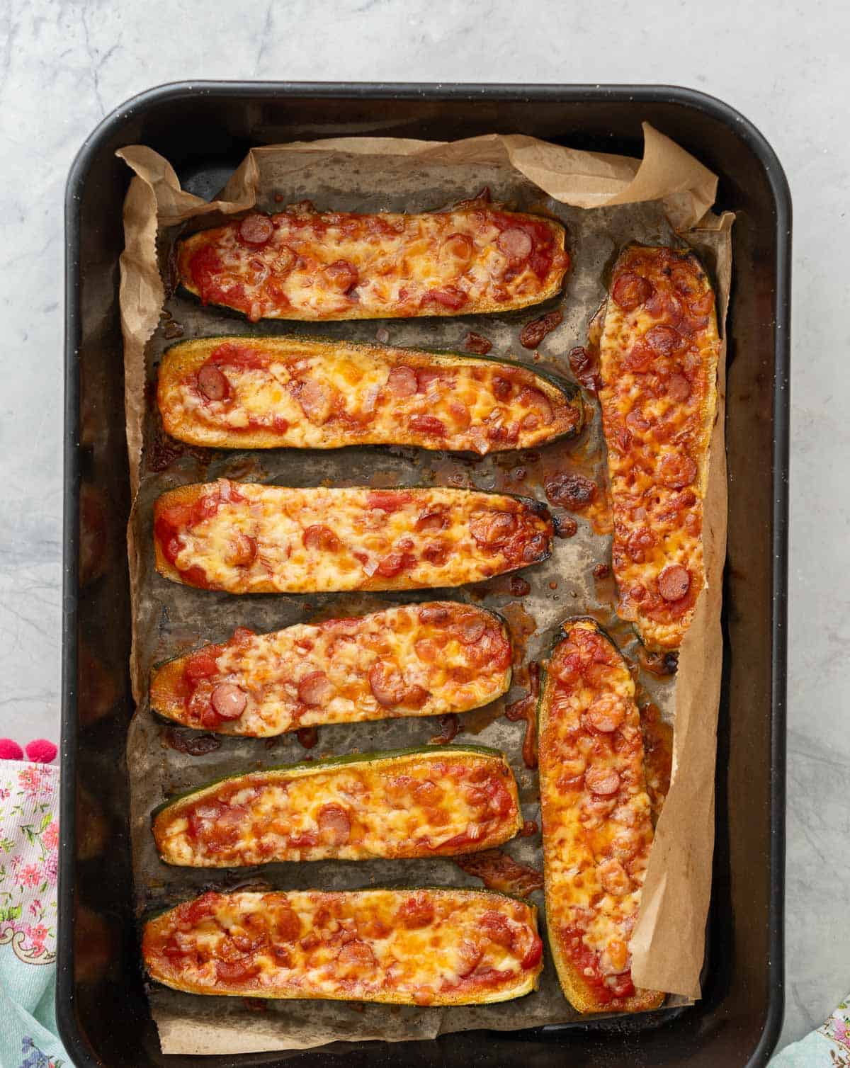 Cooked zucchini pizza boats in baking tray topped with Pizza Sauce, red onion, salami and mozzarella melted over the top.