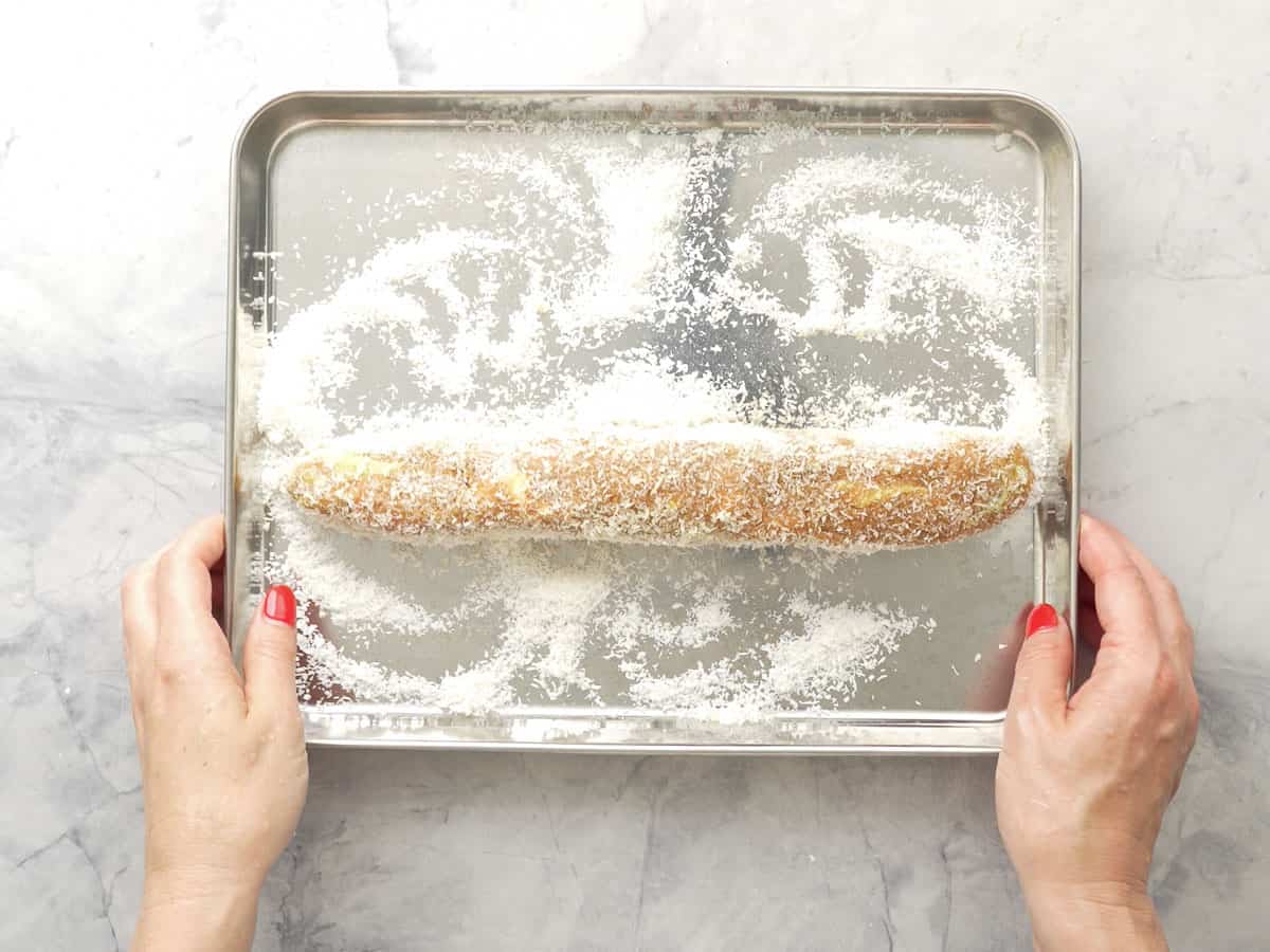 GF lolly cake log rolling in metal dish with desiccated coconut covering it.