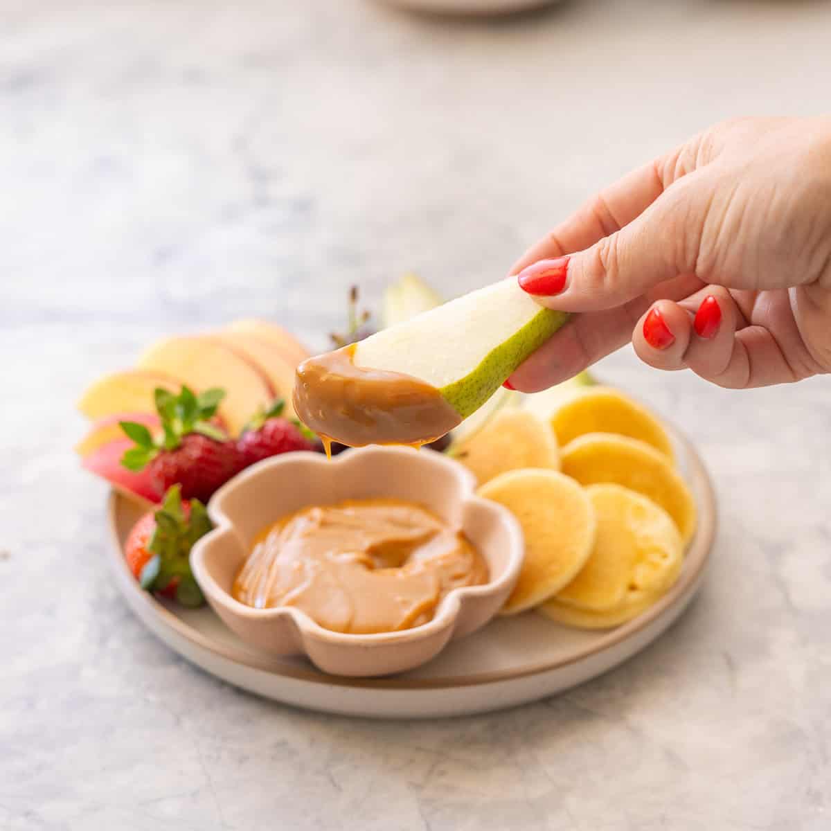 Hand holding a slice of pear dipped into Condensed milk caramel in small ramekin on plate with sliced apples, pears, grapes, strawberries and mini pancakes.