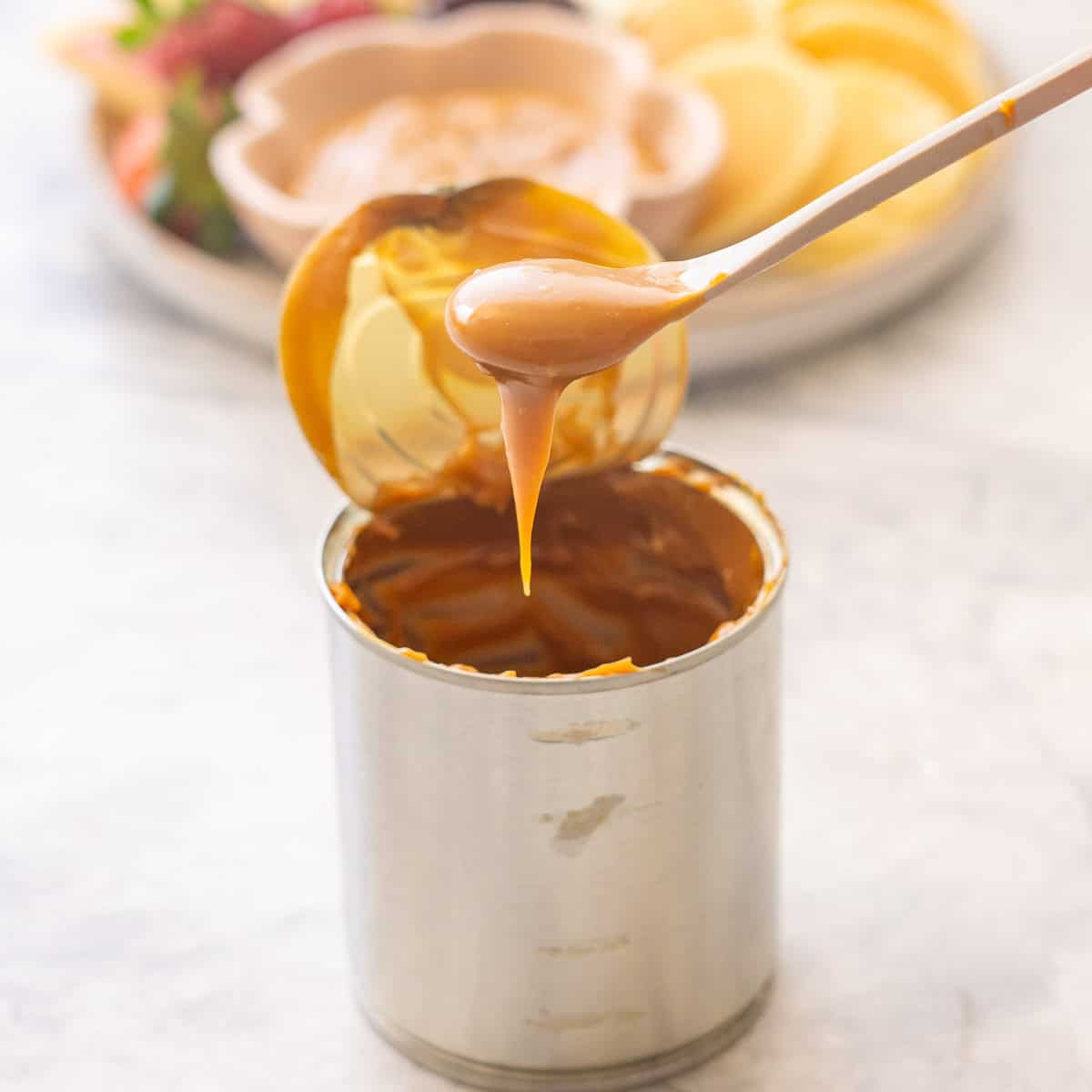 Spoonful of Condensed milk caramel being scooped out of can with lid taken off. Plate of Condensed milk caramel in small ramekin on plate with sliced apples, pears, grapes, strawberries and mini pancakes blurred in background.