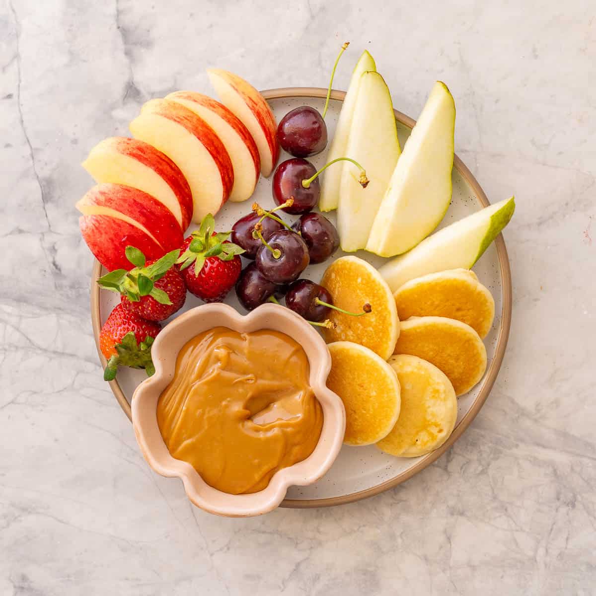 Condensed milk caramel in small ramekin on plate with sliced apples, pears, grapes, strawberries and mini pancakes.