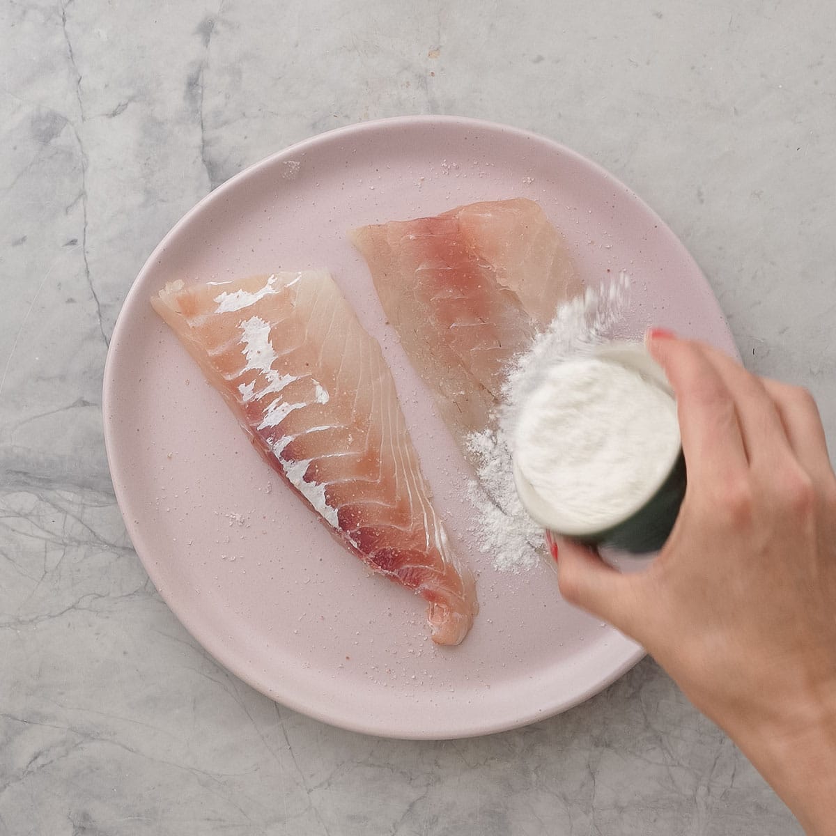 Hand holding a small ramekin of flour sprinkling it on top of two pieces of fish on a blush pink plate.