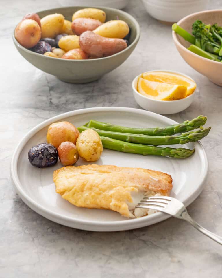 Piece of pan fried fish and potatoes with asparagus on dinner plate, bowl of seasoned potatoes and bowl of asparagus and broccolini with small bowl of lemon slices on bench top.