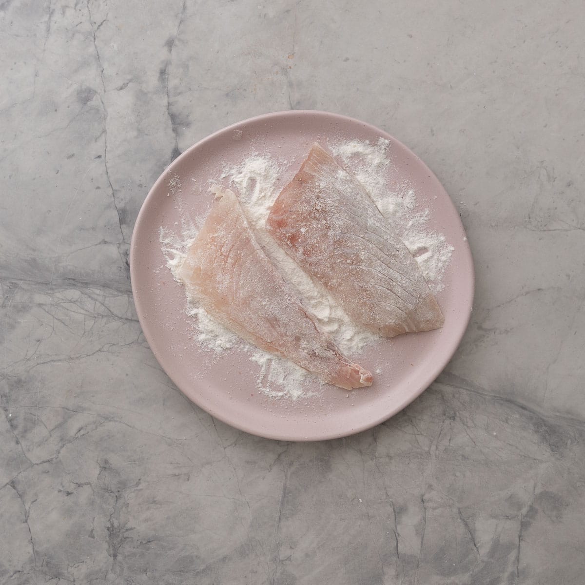 Two pieces of fish on a blush pink plate coated in flour.