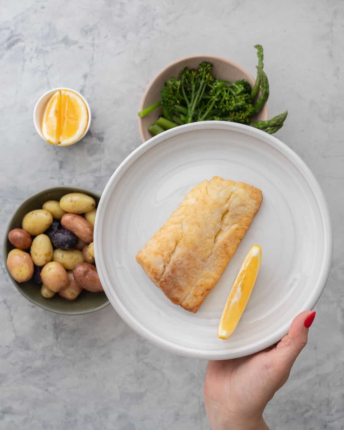 Hand holding a plate with a piece of pan fried fish and slice of lemon up to camera with bowl of seasoned potatoes and bowl of asparagus and broccolini with small bowl of lemon slices on bench top.