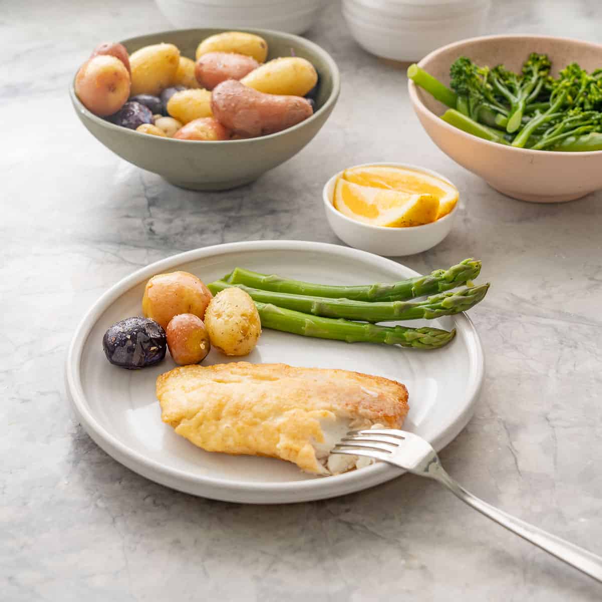 Piece of pan fried fish and potatoes with asparagus on dinner plate, bowl of seasoned potatoes and bowl of asparagus and broccolini with small bowl of lemon slices on bench top.