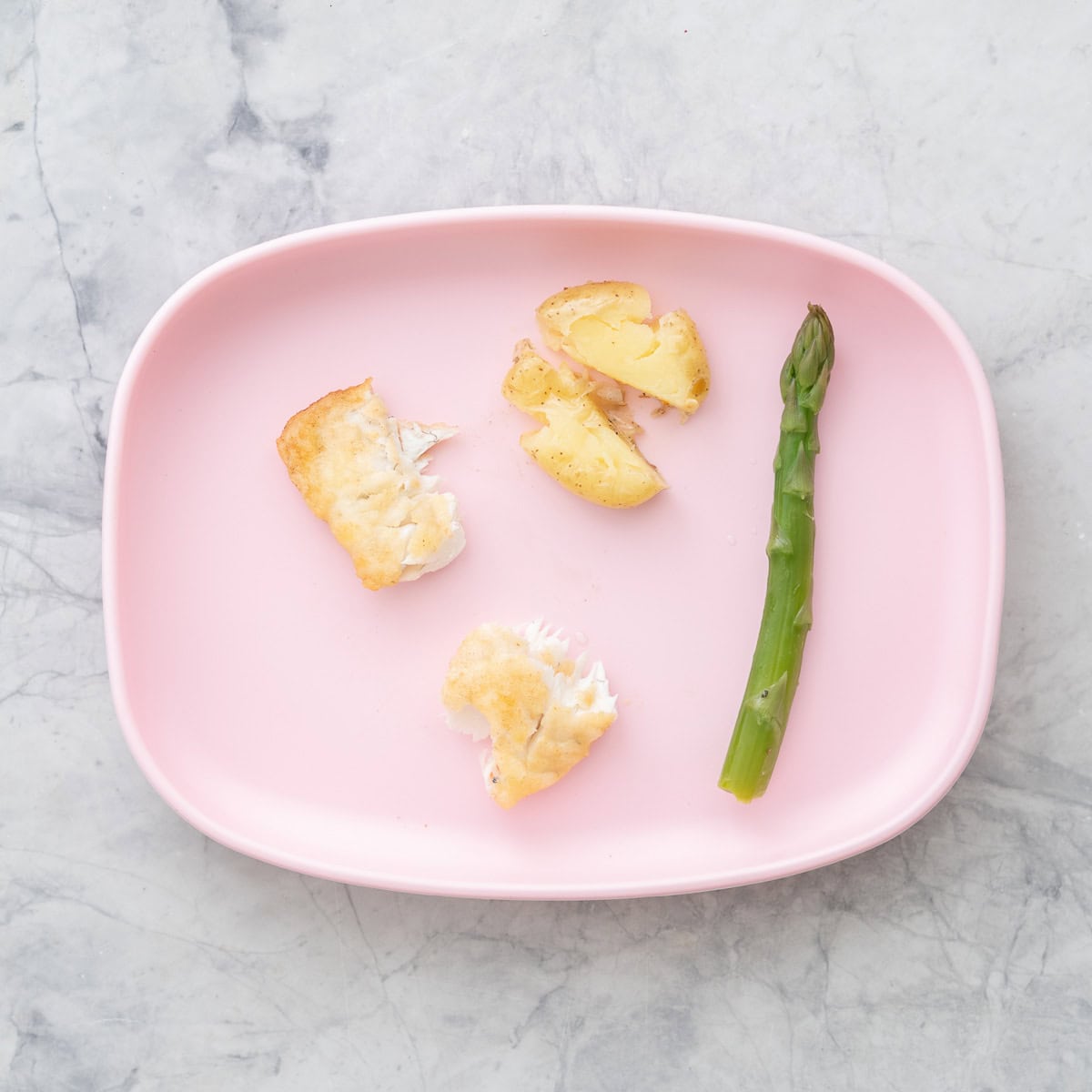 Pink silicone baby plate with a piece of asparagus, pan fried fish in two small pieces and a potato cut in half.