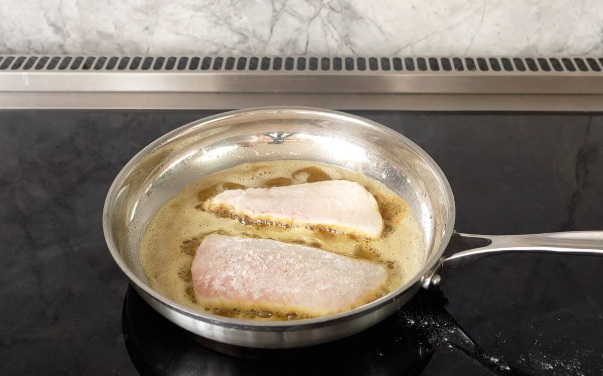 Two pieces of pan fried fish in pan of melted butter on stove top.