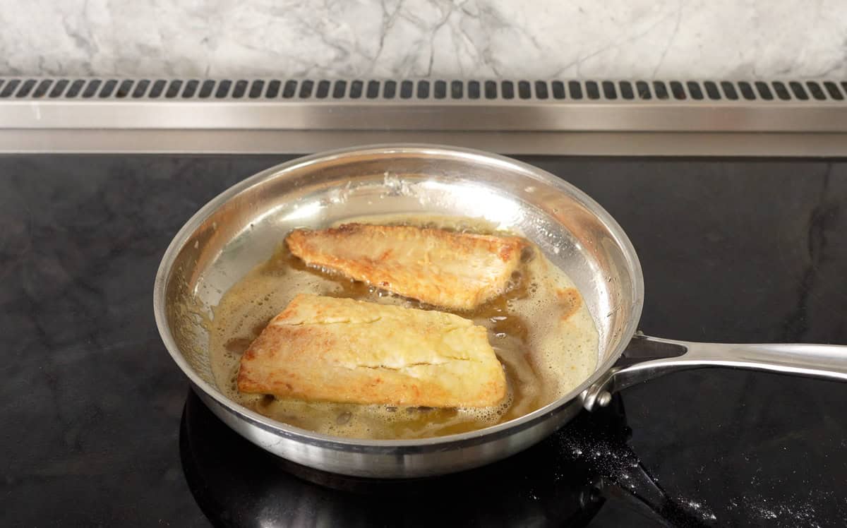 Two pieces of pan fried fish in pan of melted butter on stove top, cooked side up showing golden cook on fish.