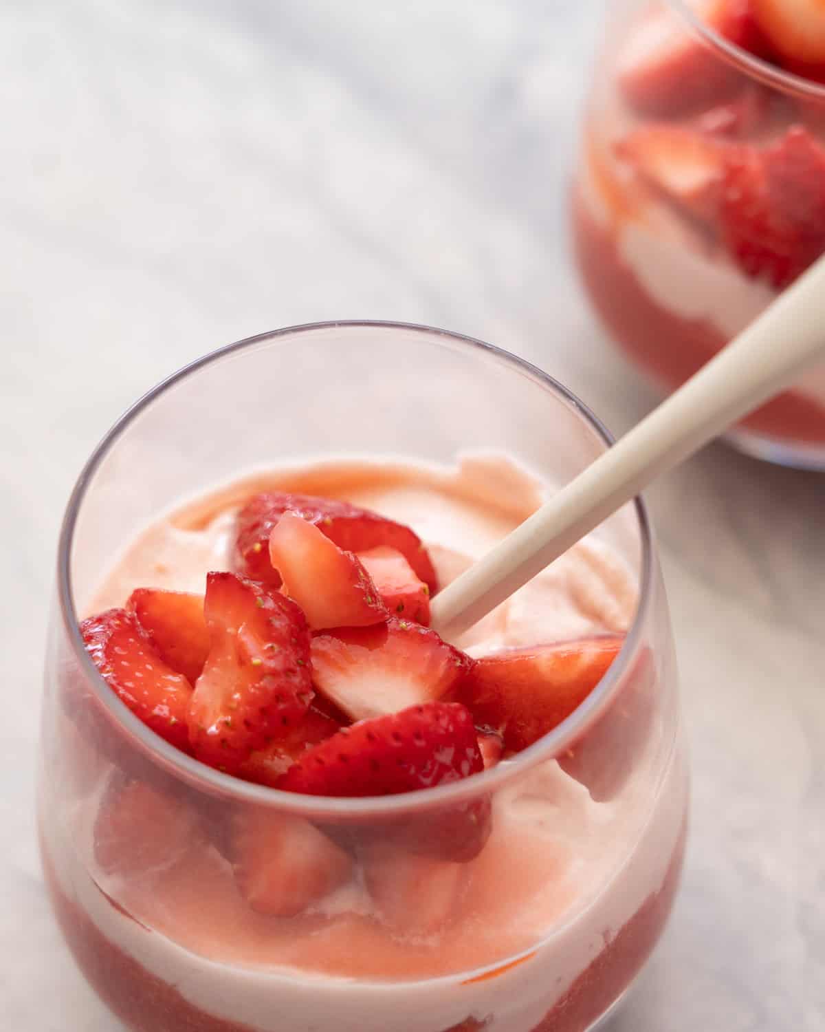 Glass with spoon inside of layered strawberry puree at base of glass, cream mixture layered on top and sliced macerated strawberries on top of cream.