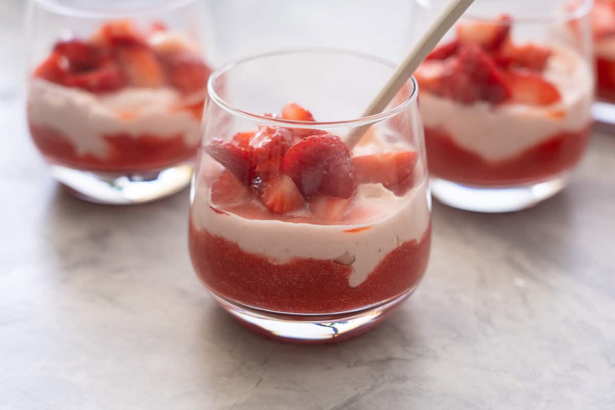 Glass with spoon inside of layered strawberry puree at base of glass, cream mixture layered on top and sliced macerated strawberries on top of cream.