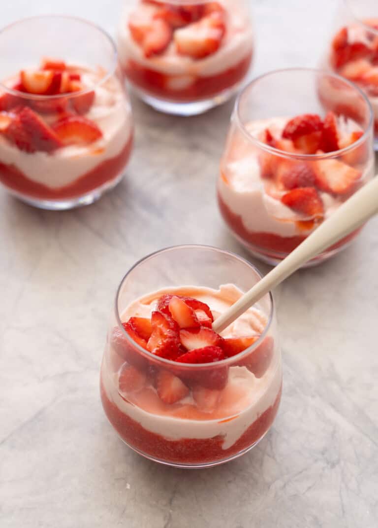 Six glasses, one with a tall spoon inside with layered strawberry puree at base of glass, cream mixture layered on top and sliced macerated strawberries on top of cream.