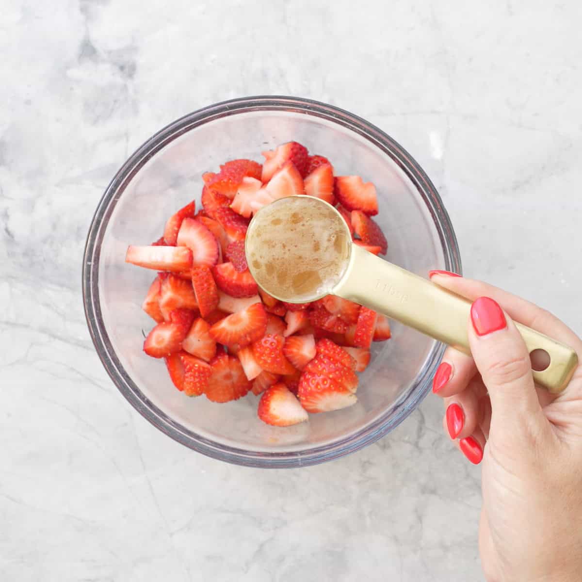 Strawberries cut in halves in a glass bowl on bench top with hand holding a measuring spoon of honey above the bowl.