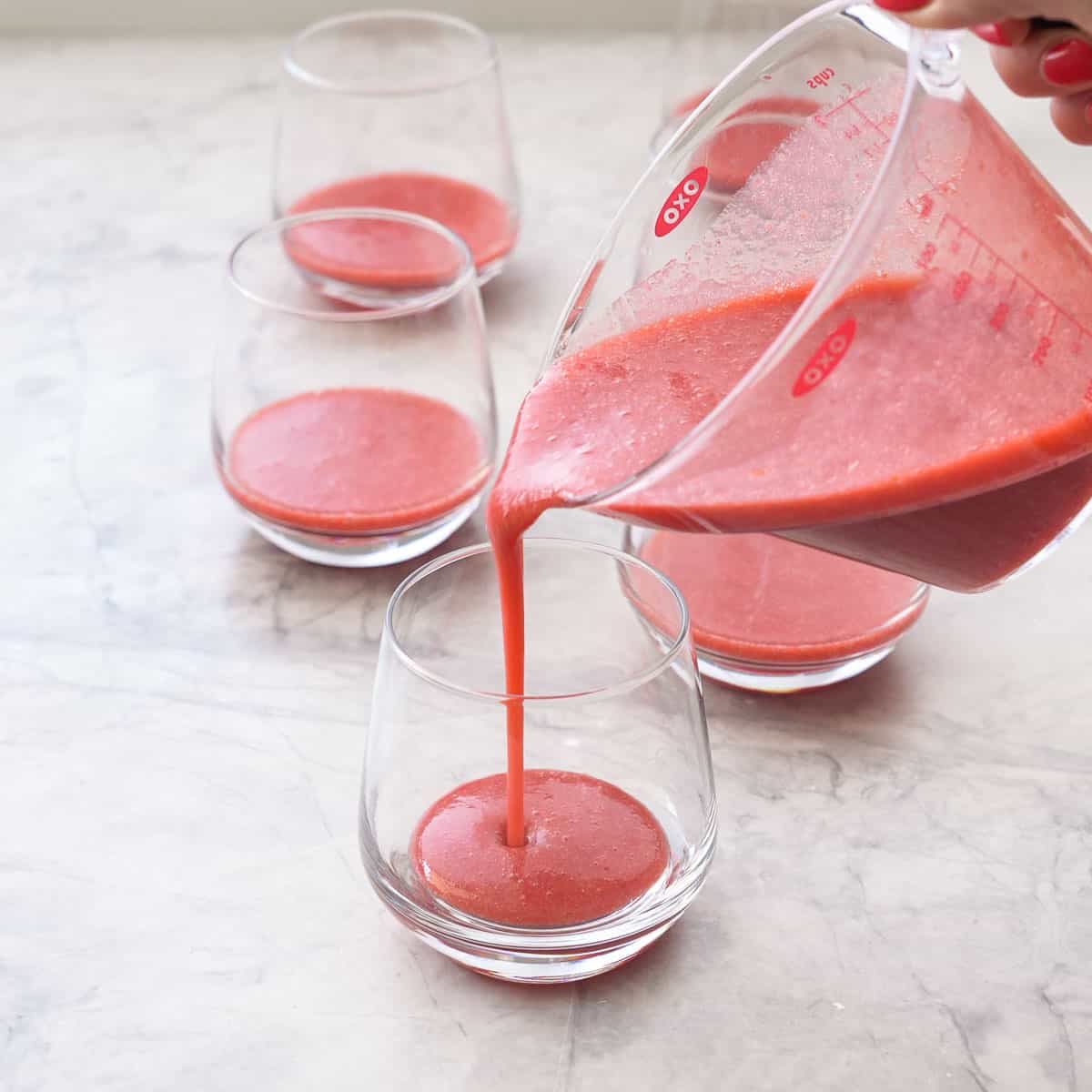 Strawberry puree in a jug being poured into the bottom of a glass with four other glasses on bench top.