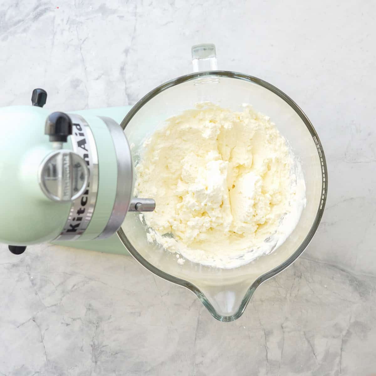 Cream, icing sugar and vanilla extract whipped together in a stand mixer glass bowl on benchtop.