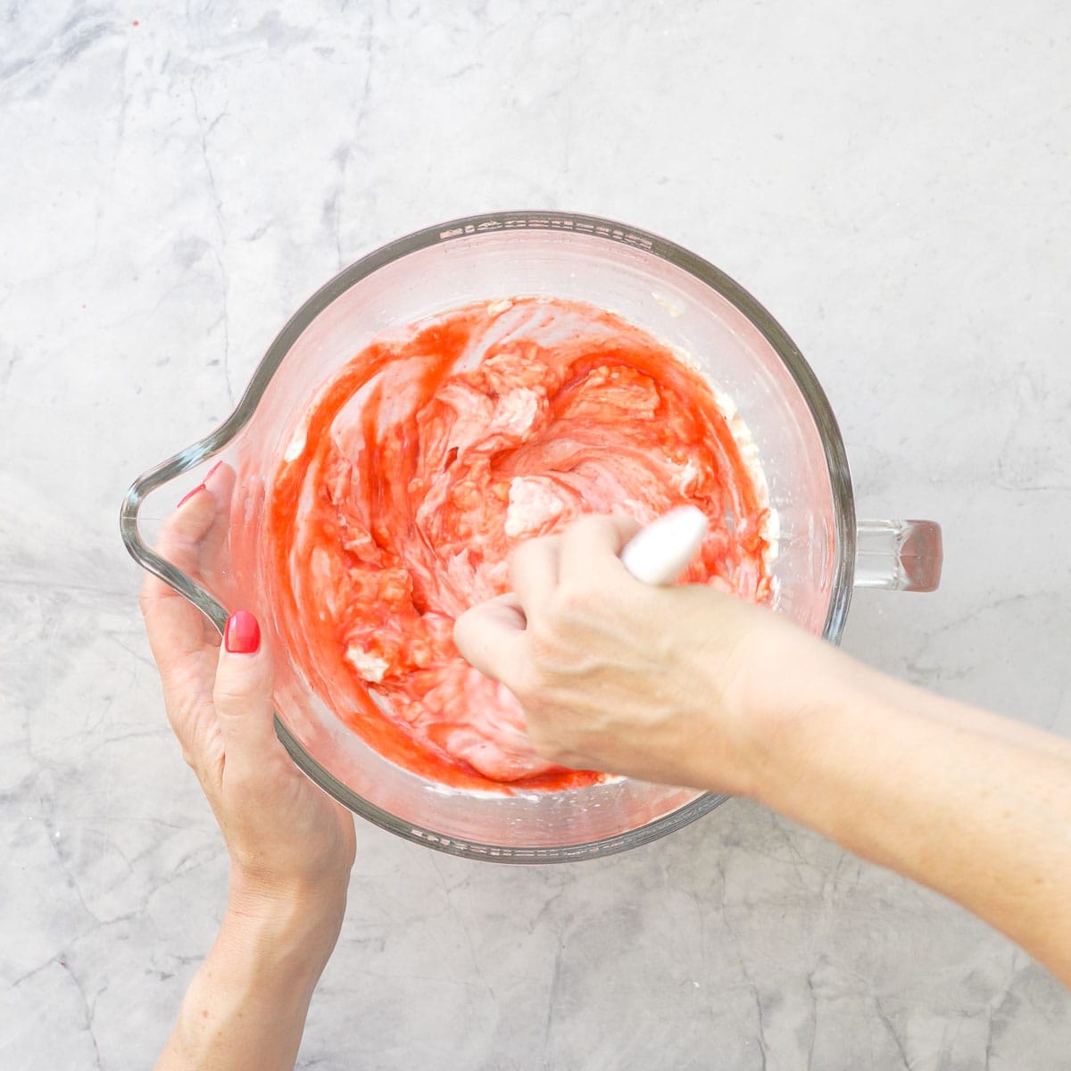 Hand holding a spatula mixing together strawberry puree mixture and whipped cream in a glass bowl on bench top.