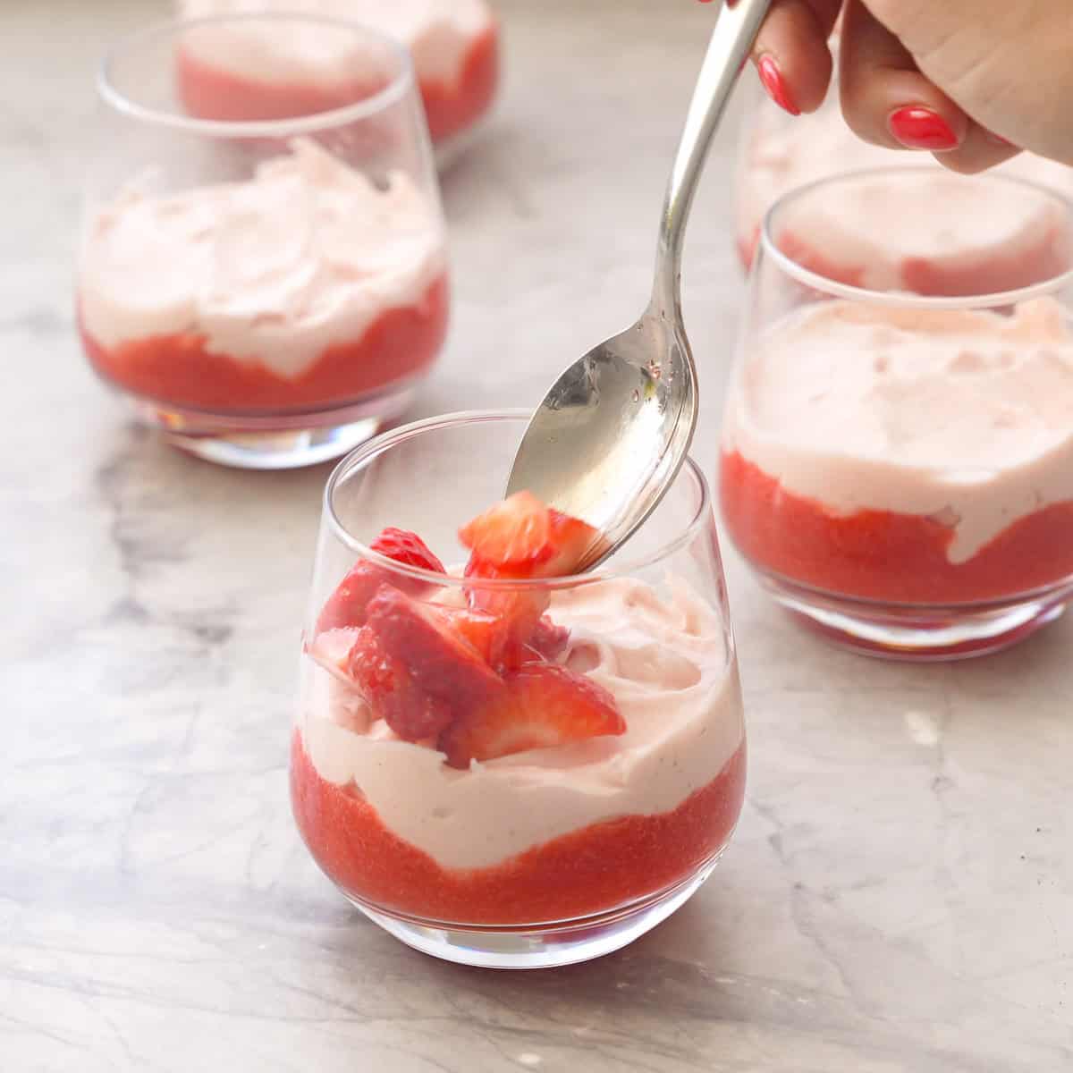 Spoon placing sliced strawberries on top of layered strawberries and cream in glass.