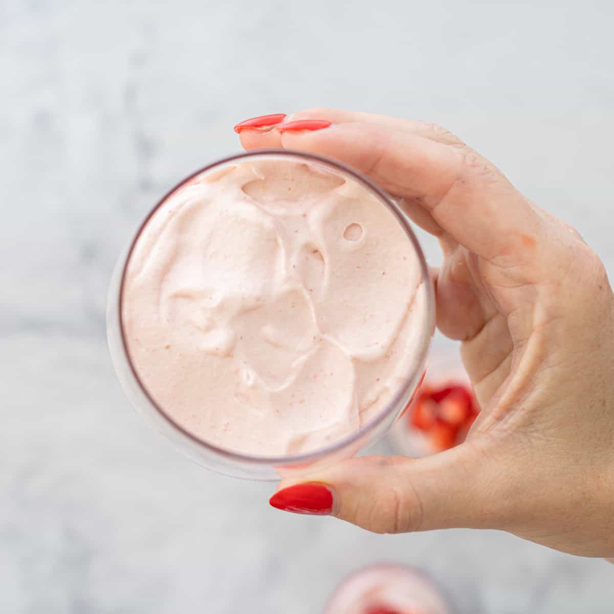 Hand holding a glass up to camera of layered strawberry puree with cream mixture on top.