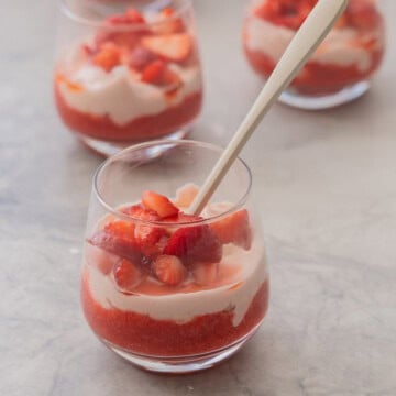 Glass with spoon inside of layered strawberry puree at base of glass, cream mixture layered on top and sliced macerated strawberries on top of cream.