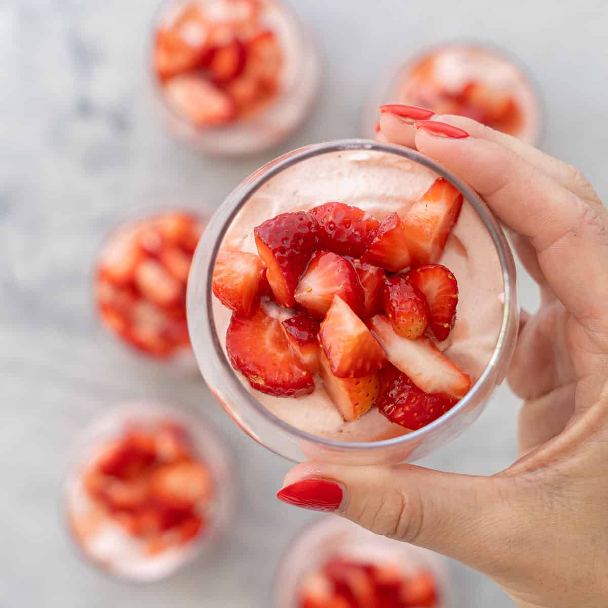 Hand holding a glass with layered strawberry puree at base of glass, cream mixture layered on top and sliced macerated strawberries on top of cream.