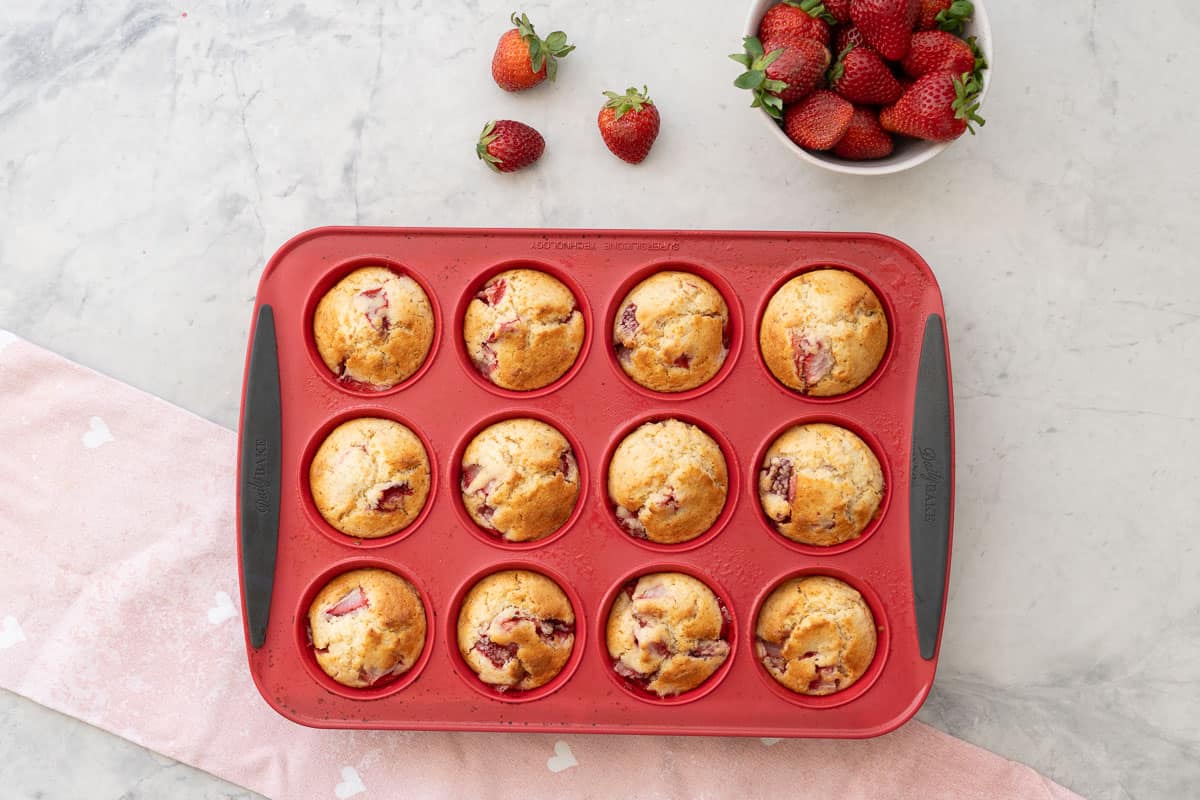 A dozen Strawberry muffins in 12 hole muffin tray on bench top with a light pink tea towel and small bowl of fresh strawberries.