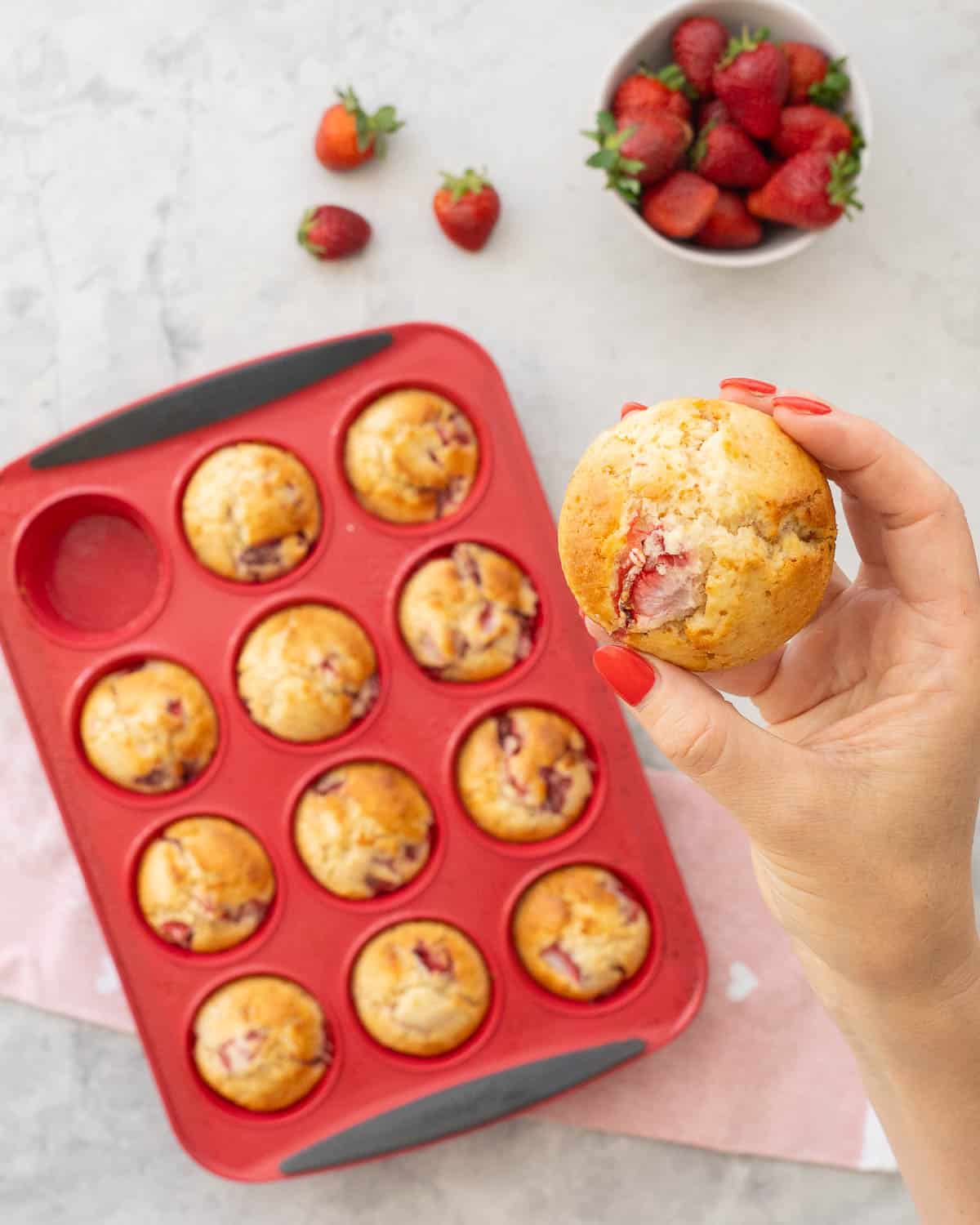 Hand holding a strawberry muffin up to camera with plate below with five muffins and 12 hole muffin tray with muffins inside on bench top with light pink tea towel.