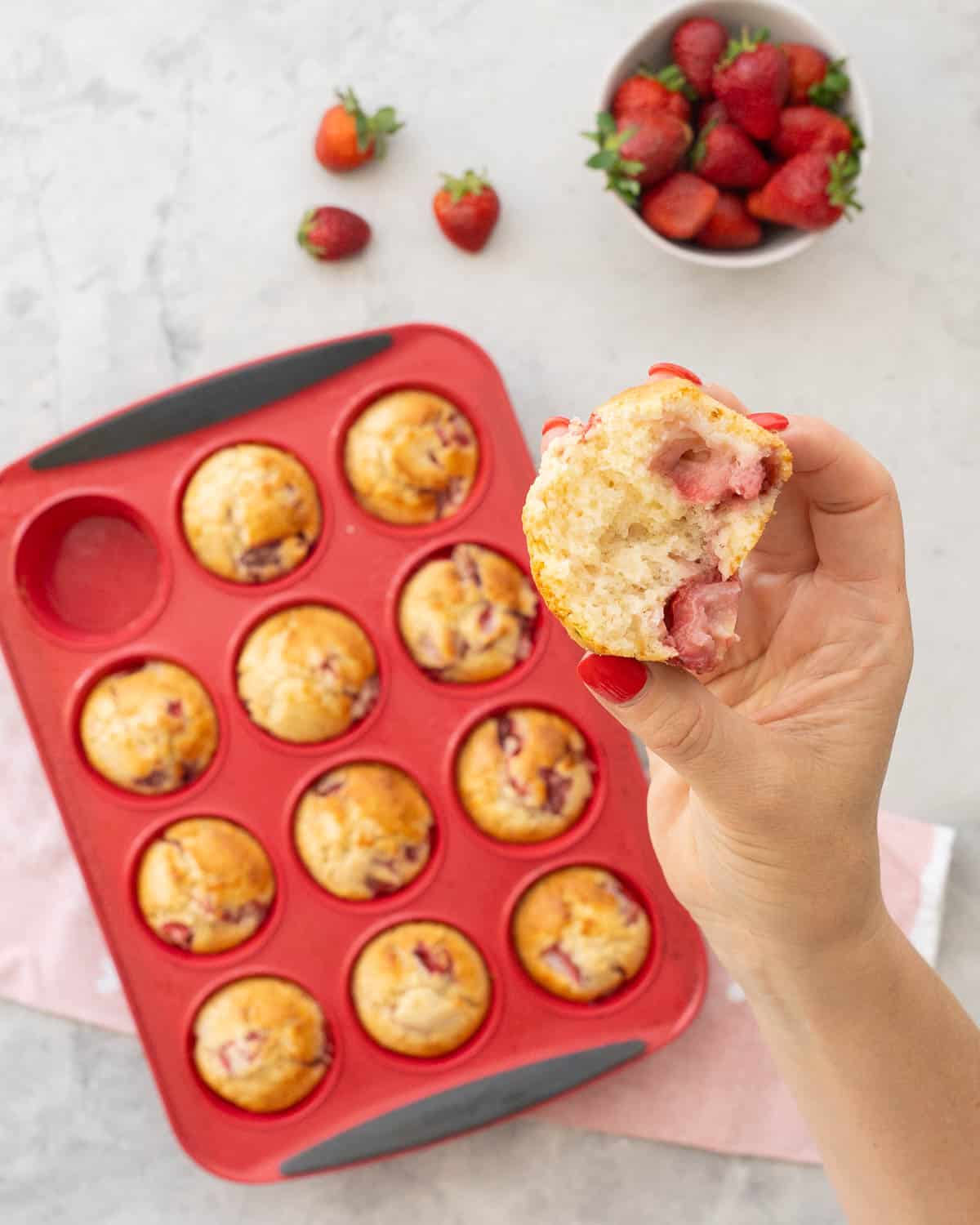 Hand holding a strawberry muffin broken in half up to camera with plate below with five muffins and 12 hole muffin tray with muffins inside on bench top with light pink tea towel.