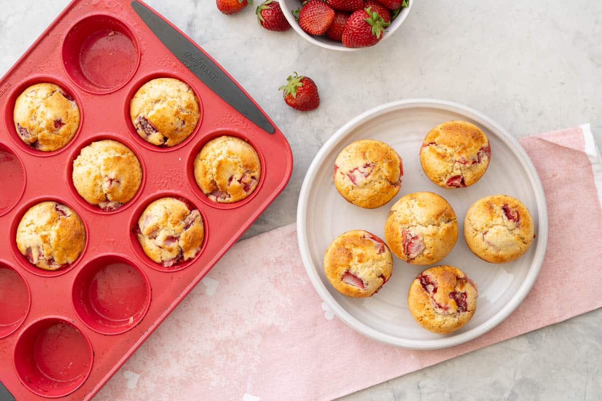 Five muffins on a plate and muffin tray with muffins inside and a bowl of strawberries on bench top with light pink tea towel.