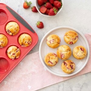 Five muffins on a plate and muffin tray with muffins inside and a bowl of strawberries on bench top with light pink tea towel.