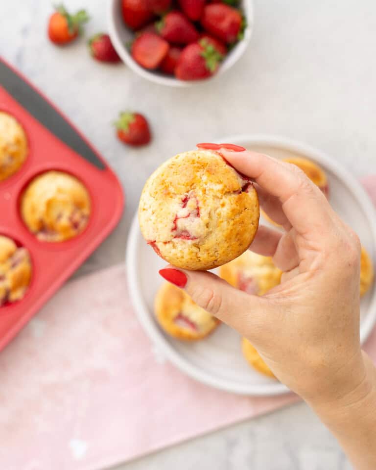 Hand holding a strawberry muffin up to camera with plate below with five muffins and muffin tray with muffins inside on bench top with light pink tea towel.