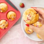 Hand holding a strawberry muffin up to camera with plate below with five muffins and muffin tray with muffins inside on bench top with light pink tea towel.