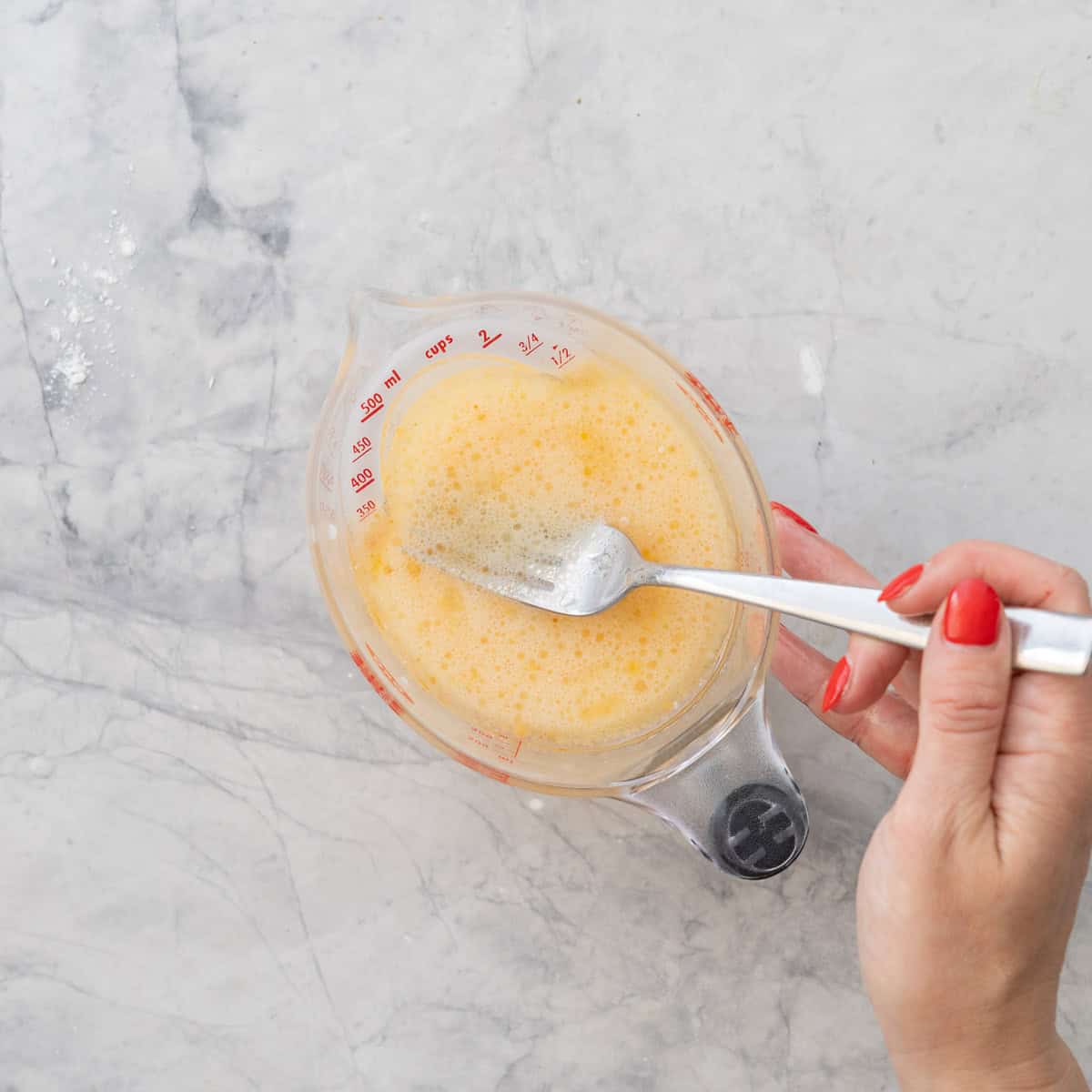 Small jug on bench top with oil, egg, milk and vanilla whisked together with hand holding a fork inside. 