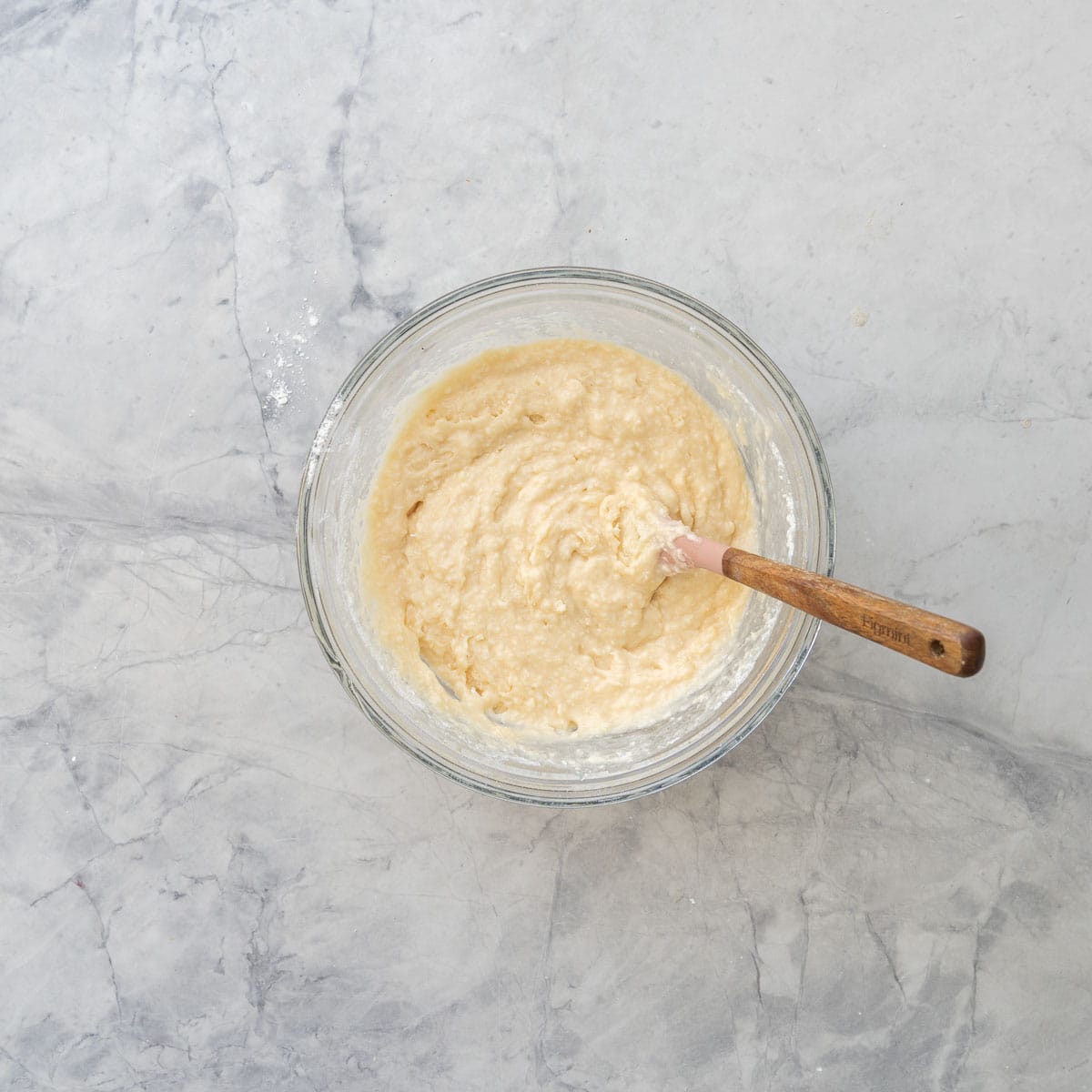 Wet and dry ingredients mixed together in a glass bowl with spatula inside on bench top.