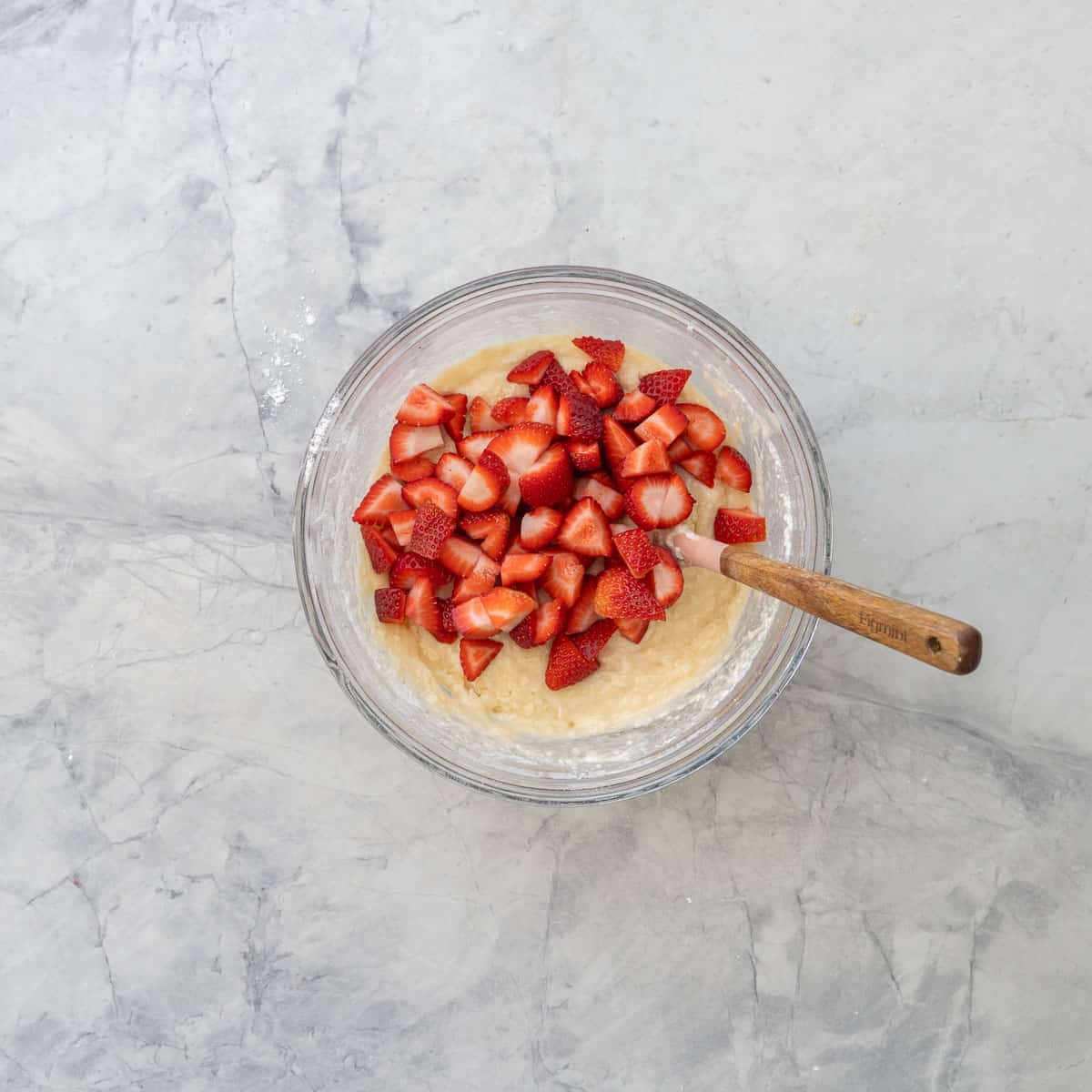 Cut strawberries on top of muffin mixture in glass bowl on benchtop.