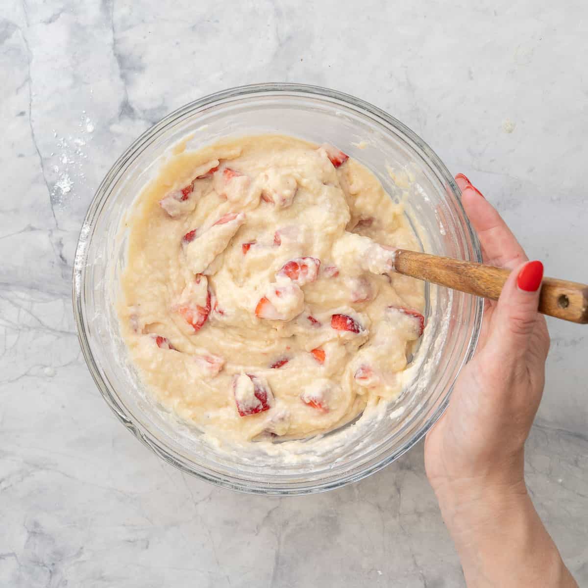 Strawberries mixed into strawberry muffin mixture with hand holding a spatula in glass bowl on bench top.