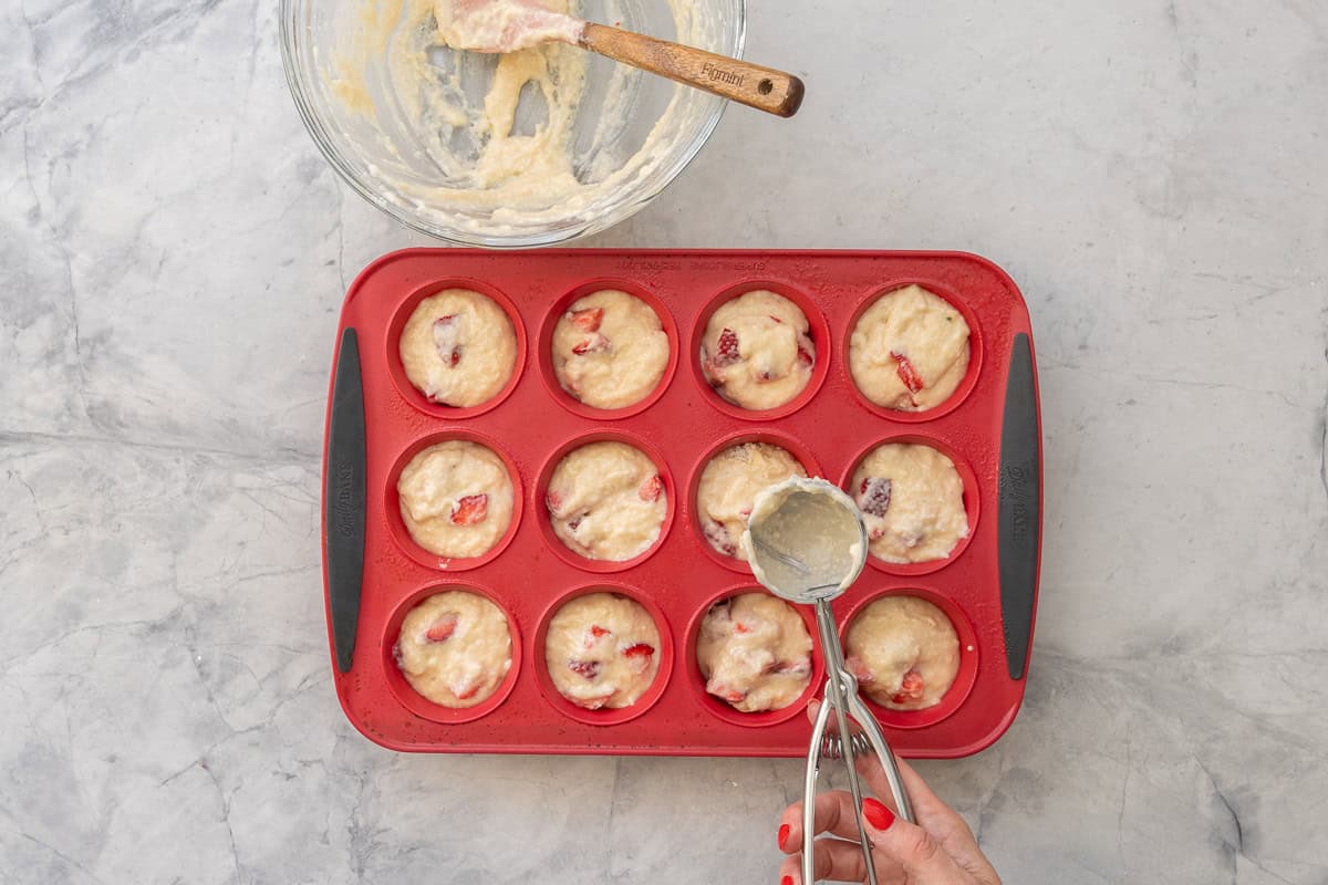 Hand holding a cookie scooper with Strawberry muffin batter residue, bowl with batter and spatula inside and a 12 hole muffin tray filled with scoops of strawberry muffin batter.